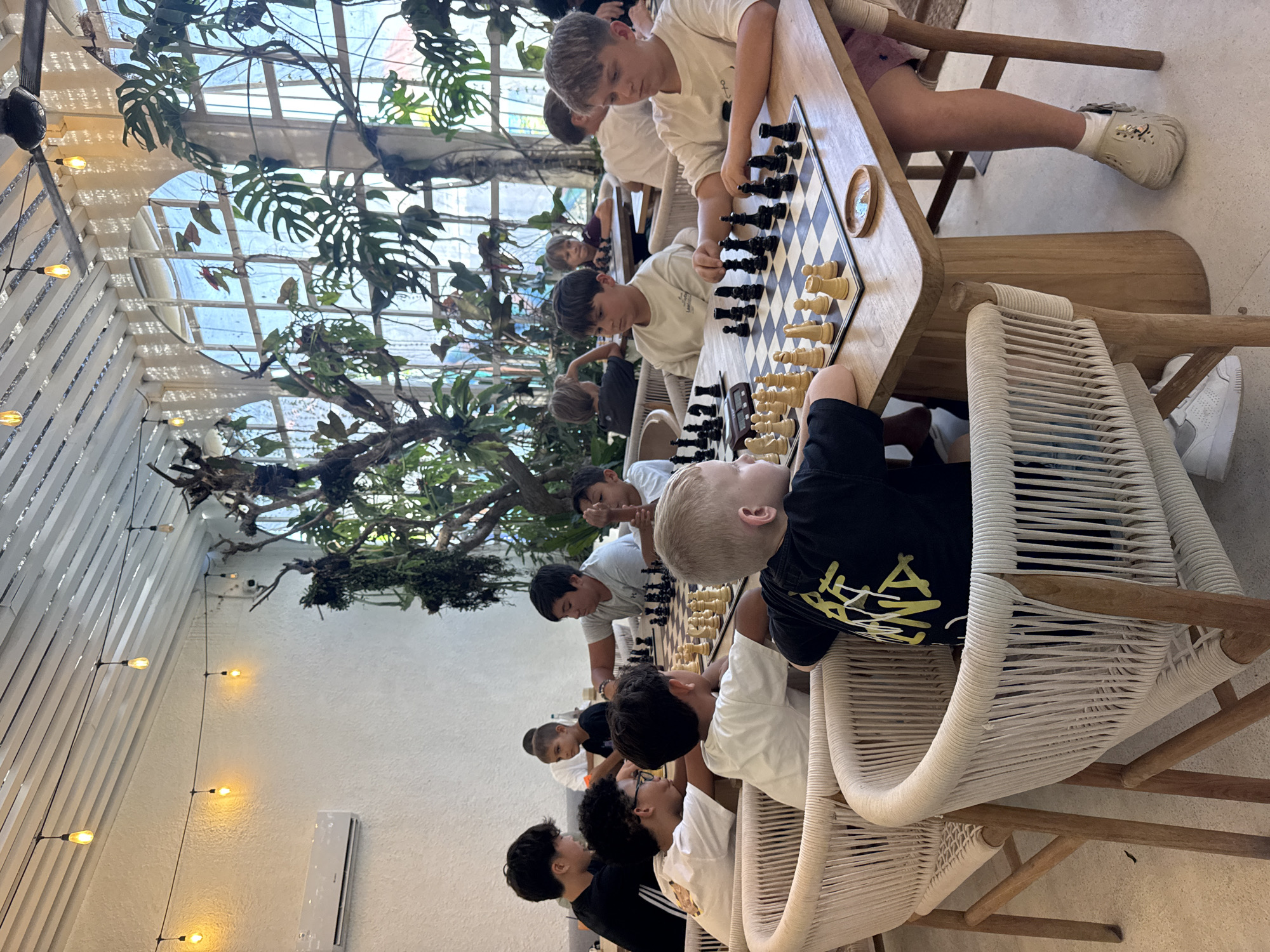 A group of young people play chess at wooden tables in an indoor setting. The tables and chairs are crafted from light-colored wood, with the chairs featuring woven rope seats and backs. The location appears to be a cafe or restaurant, possibly in North Kuta, Indonesia, given the tropical foliage visible behind a large window. The interior decor includes a wall lined with string lights. The children are focused on their games.