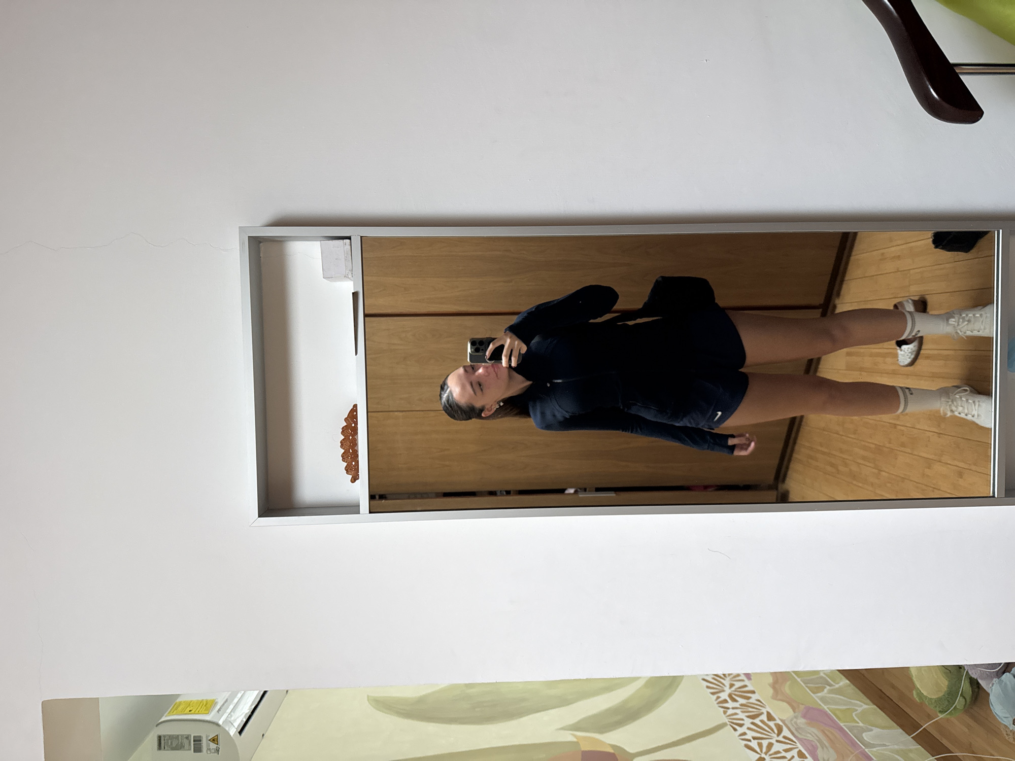 A full-length mirror reflecting a young woman taking a selfie is mounted on a wall. The woman is wearing a dark-colored athletic outfit consisting of shorts and a zip-up jacket, paired with white sneakers and socks. A black handbag hangs on her shoulder. The mirror reflects a bedroom interior, including a wooden wardrobe, and a wooden floor. A white wall with a noticeable crack, a coat hanger, an air conditioner, and a portion of a decorative painting are visible around the mirror. The location is an indoor space in San Bernardino Tlaxcalancingo, Mexico.