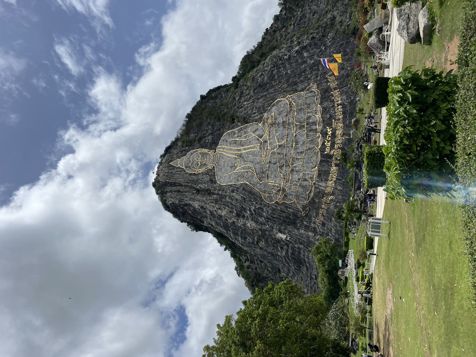 The image shows a large cliff face in Na Chom Thian, Thailand, carved with a golden depiction of Buddha. The Buddha image is seated in meditation, outlined in gold against the grey rock. Below the Buddha, there is golden text in Thai script. At the base of the cliff, there is a landscaped park area with grass, trees, and some people walking and sitting. Two flags, one Thai flag and one yellow flag, are visible on flagpoles near the cliff. The sky above is partly cloudy with patches of blue.