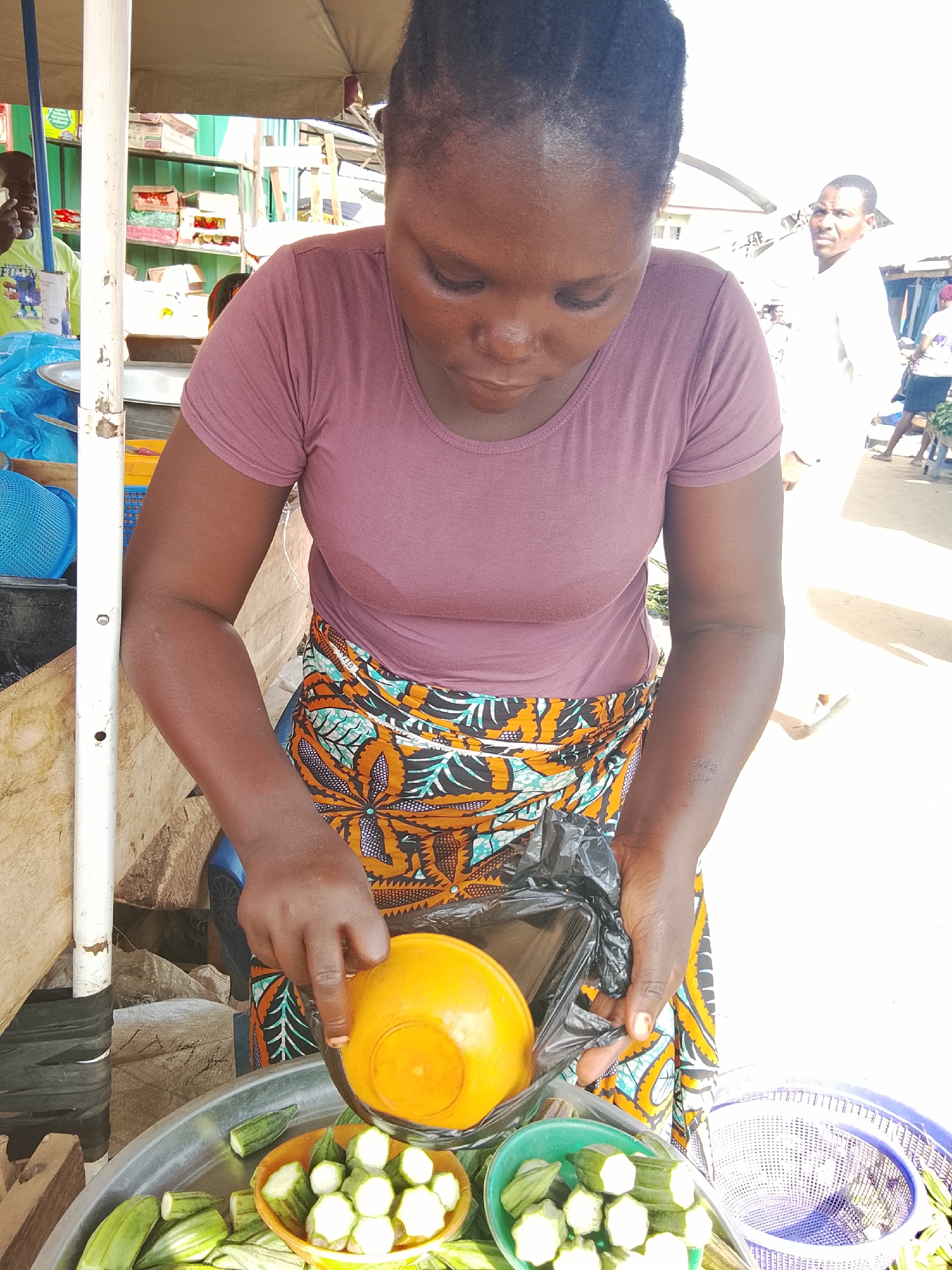 In Akwanga, Nigeria, a Black woman with dark braided hair is shown from the chest up, wearing a dusty rose t-shirt and a brightly colored African print skirt. She is looking down at a transaction she is conducting. Her right hand holds a black plastic bag and an orange bowl containing chopped okra. Her left hand is touching the okra in the bowl. In the foreground, a large metal platter and two smaller bowls are filled with okra. In the background, a man in a white shirt is visible. The scene takes place outdoors, possibly at a market stall, under a canopy.