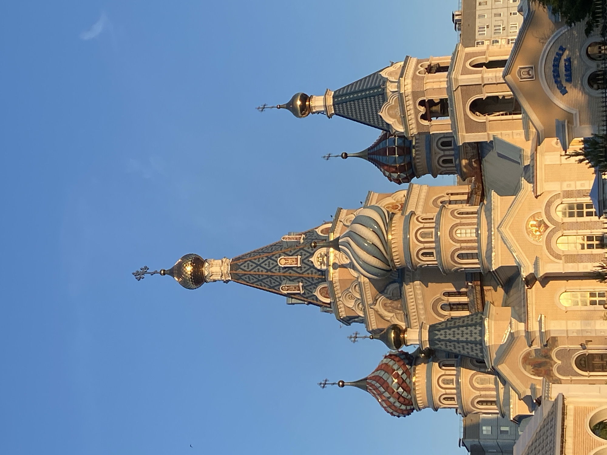 This image captures the Cathedral of the Archangel Michael in Izhevsk, Russia, under clear blue skies. The prominent multi-domed Eastern Orthodox church is illuminated by warm, golden light, indicating either sunrise or late afternoon. The central and tallest spire features a conical roof composed of blue and light-colored diamond-patterned tiles, capped by a gleaming golden onion dome and a cross. To the left of the central spire, a smaller dome exhibits a reddish-brown and light-colored scale-like pattern. To the right, another dome displays a distinctive blue and white striped, twisted pattern. Several other smaller golden onion domes, each topped with a cross, are integrated into the cathedral's structure. The building's facade is primarily light yellow/beige, adorned with numerous arched windows, decorative elements, and small religious icons visible in niches. A bell tower containing visible bells is located on the right side of the edifice, topped with a blue patterned roof and a golden onion dome. A sign with Cyrillic text reading "КАФЕДРАЛЬНЫЙ СОБОР" (Cathedral) is visible on the lower right portion of the building. In the background, modern multi-story residential buildings are partially visible. Dark green foliage is present along the bottom edge of the frame. The sky is a uniform light blue, with a small dark bird visible in the upper left quadrant and a faint, wispy cloud formation in the upper right. No individuals are present in the scene.