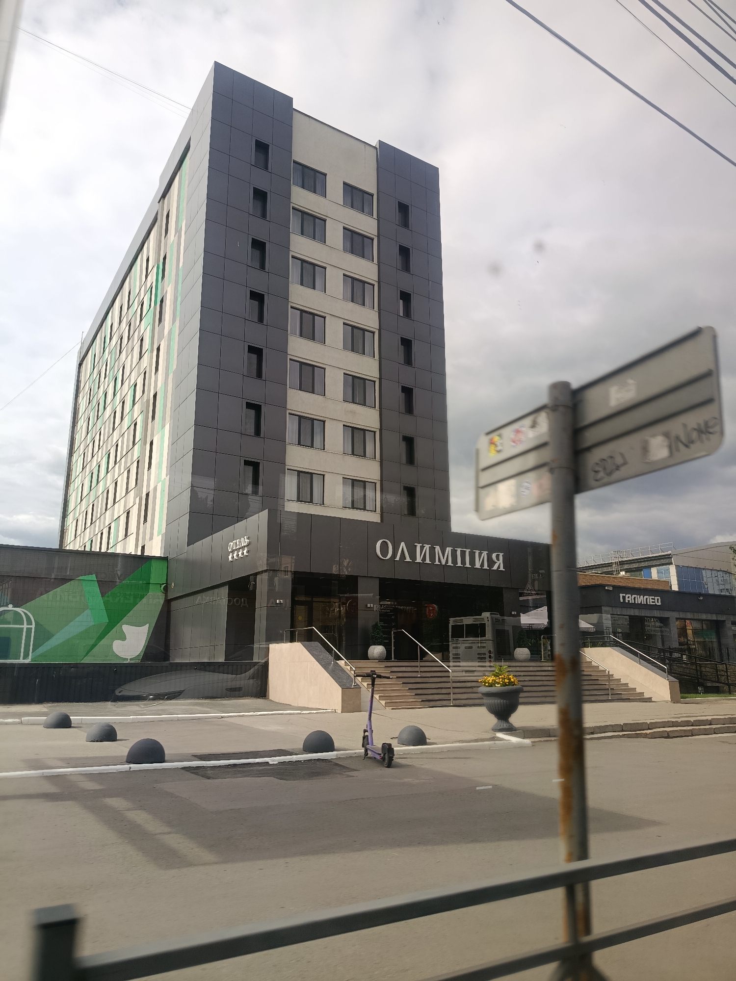The image shows the "Olimpiya" hotel in Saratov, Russia, as evidenced by the Cyrillic lettering on the building. The hotel is a multi-story modern building with a dark grey facade on the right and a facade with green and white vertical stripes on the left. The main entrance area features a set of stairs leading up to the building, with metal handrails. In front of the hotel, there is a sidewalk with several large, dark grey, spherical bollards. A purple electric scooter is parked on the sidewalk near the stairs. Further to the right, there is a signpost with a blurred informational sign attached to it. A plant in a large decorative pot is situated near the bottom of the stairs. In the background, there is another building with the Cyrillic lettering "ГАЛИЛЕО" visible. The sky is overcast with clouds. The image appears to have been taken from a vehicle, as suggested by the blur and the presence of a railing in the foreground.