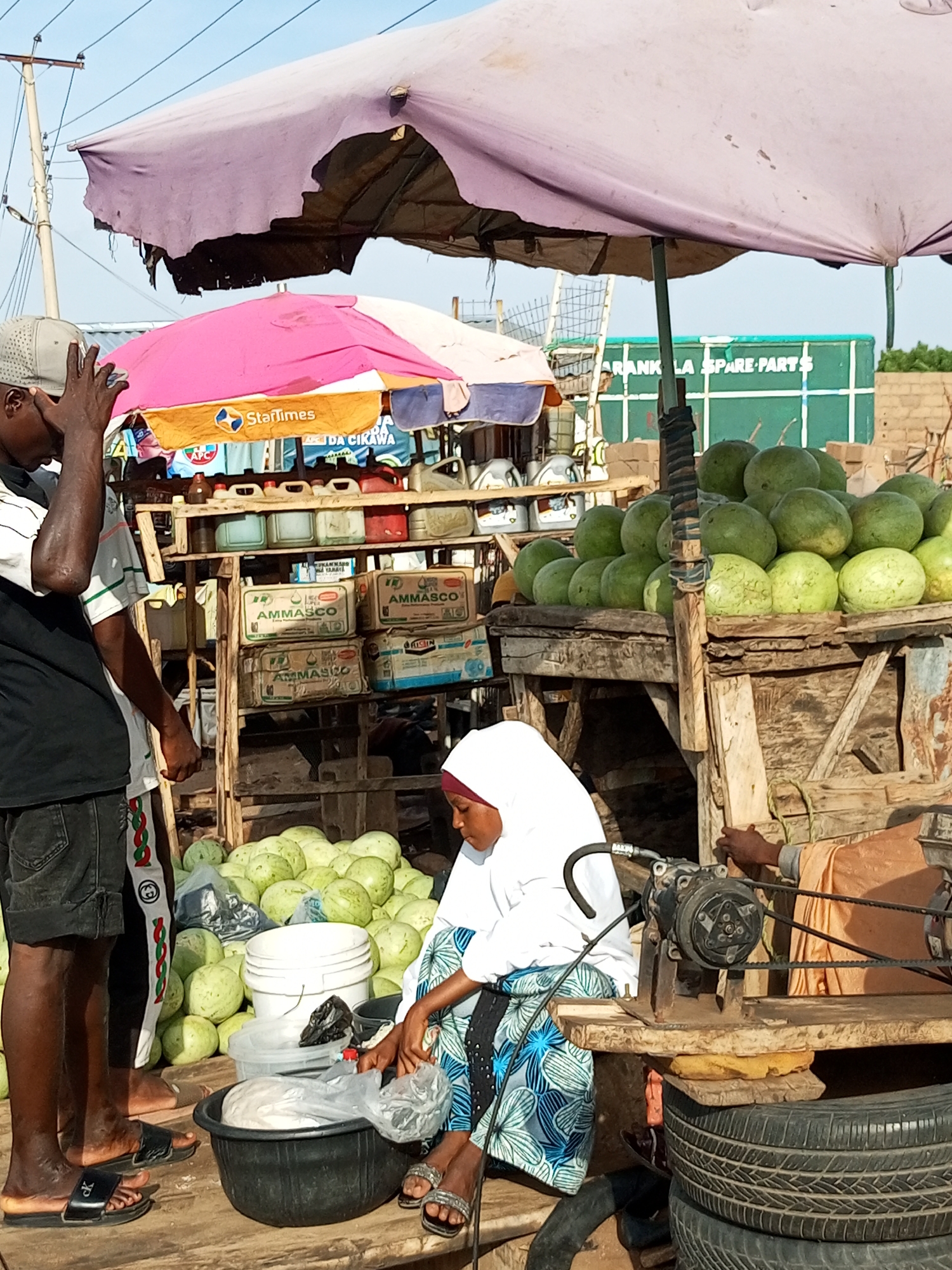 In an outdoor market scene, a group of people are gathered around stalls laden with produce and goods. The foreground features a woman wearing a white hijab and a blue and white patterned dress, seated and attending to something in a black basin. To her left, the legs of a man in shorts and sandals are visible. Behind them, a large pile of green gourds or melons is stacked on a wooden surface.
Further back, a wooden stall displays various items, including what appear to be plastic containers and canisters. Above the stall, colorful umbrellas provide shade, one pink and orange, and another larger, tattered purple one. In the background, a green structure with white lettering, possibly advertising, is visible, along with more stalls and what looks like a fence.
The time of day appears to be daytime, with bright sunlight casting shadows, suggesting fair weather. The overall setting is a bustling marketplace, likely in a tropical or sub-tropical region given the produce and outdoor conditions. The mood seems ordinary, indicative of daily commerce.
Visible text includes "StarTimes" on one of the umbrellas, and "AMMASCO" printed on several boxes stacked on the wooden stall. The green structure in the background has the words "ARANK LA SPARE PARTS" written on it.