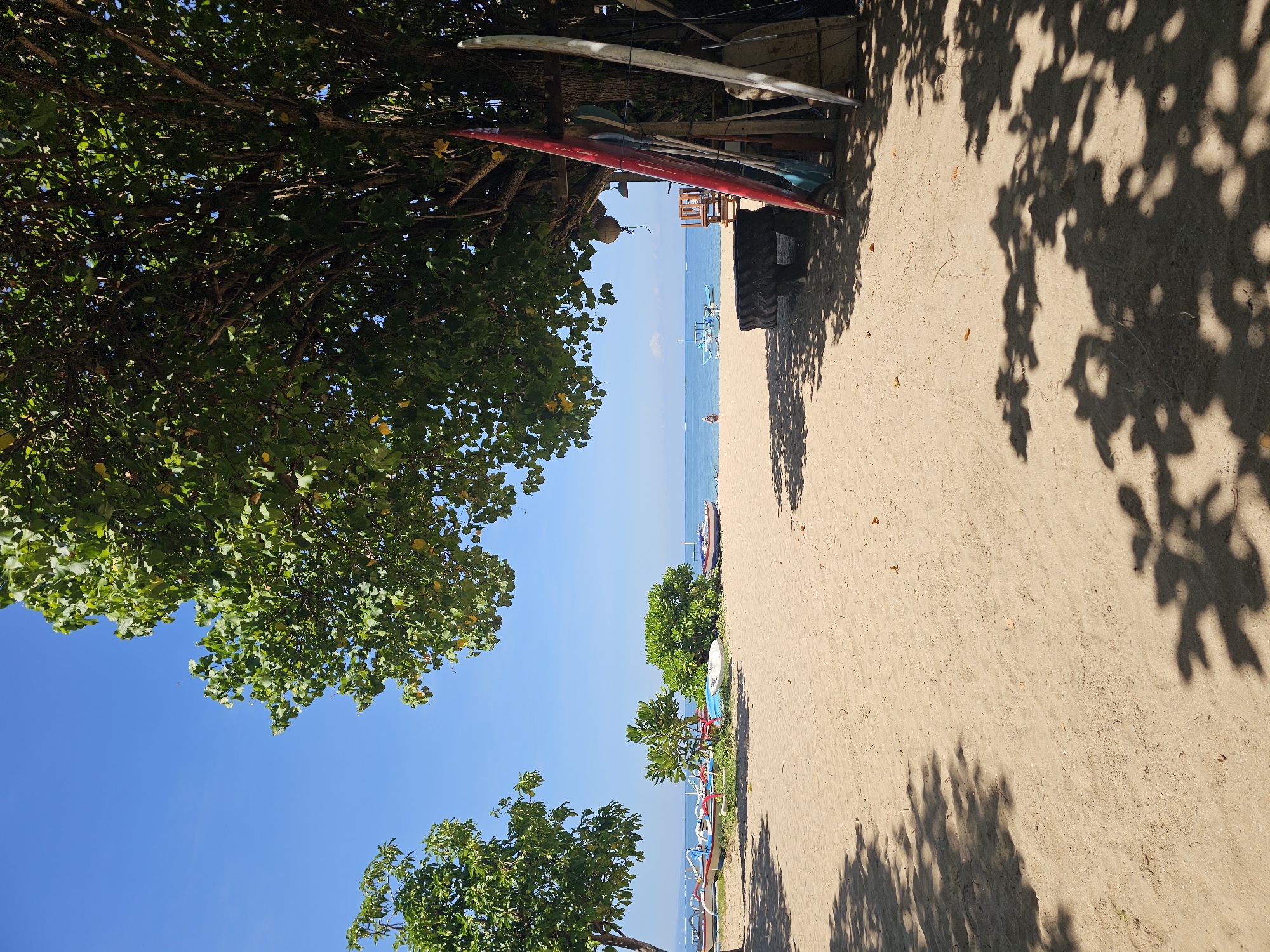 The image shows a sunny beach scene in Denpasar, Indonesia. The perspective is slightly tilted. The foreground features a sandy beach, partially covered by the shadows of trees. In the upper left corner and along the bottom of the frame, there are trees with green foliage. Various boats and surfboards are visible along the shoreline. In the distance, the blue ocean meets the clear sky. A small figure of a person can be seen walking on the beach in the middle ground. A partially visible tractor tire leans against a structure holding surfboards and other equipment.
