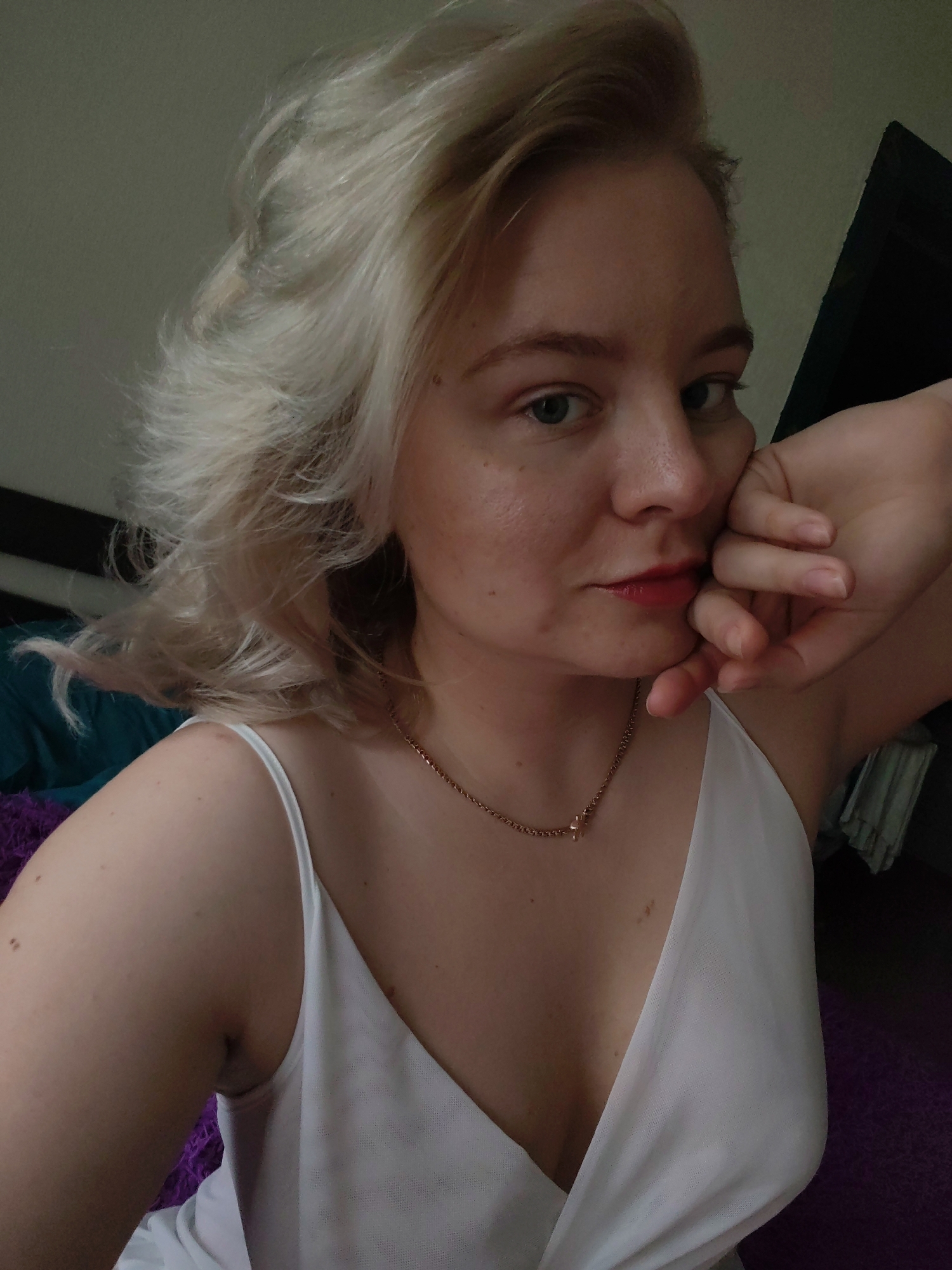 A fair-skinned woman with blonde hair is posing indoors. She is wearing a white, low-cut top and a gold necklace. Her head is resting on her hand. The background includes a dark headboard and purple and teal fabrics. The location is not discernible as being within the city of Izhevsk, Russia.