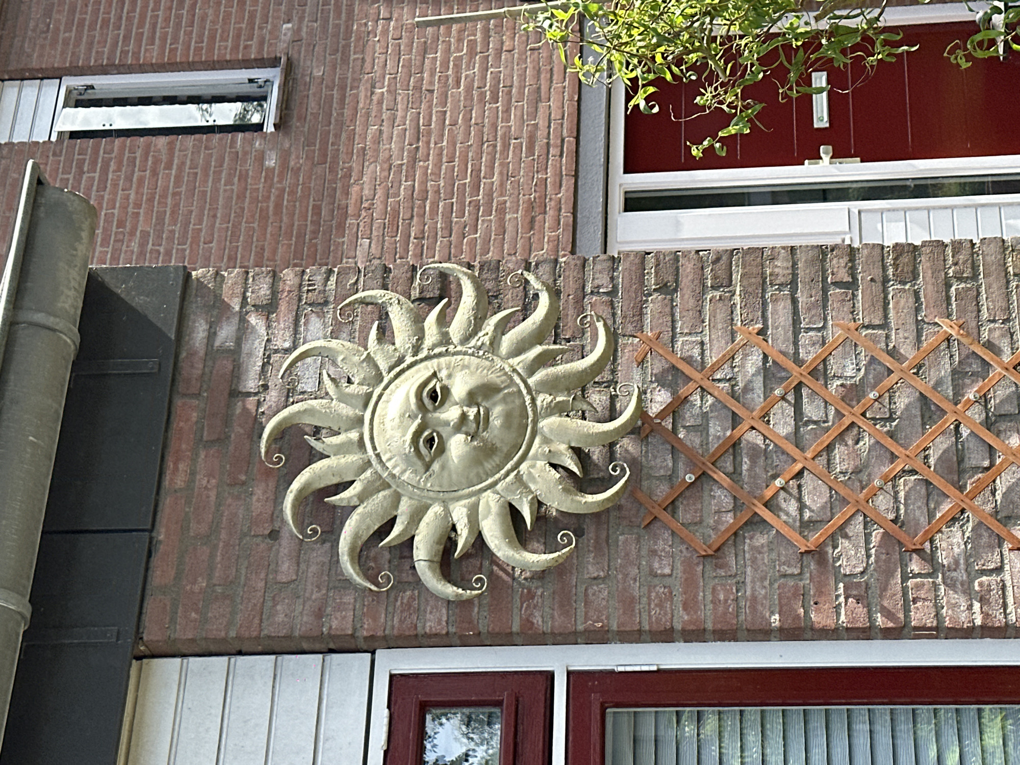 The image depicts an exterior wall of a brick building in Dronten, Netherlands. A decorative metal sun with a face is mounted on the wall. To the right of the sun is a wooden trellis. Above the brick wall are windows with white frames. Below the brick wall is a window with red frames. The scene appears to be outdoors during daylight. The environment consists of a residential building exterior in a Dutch city.