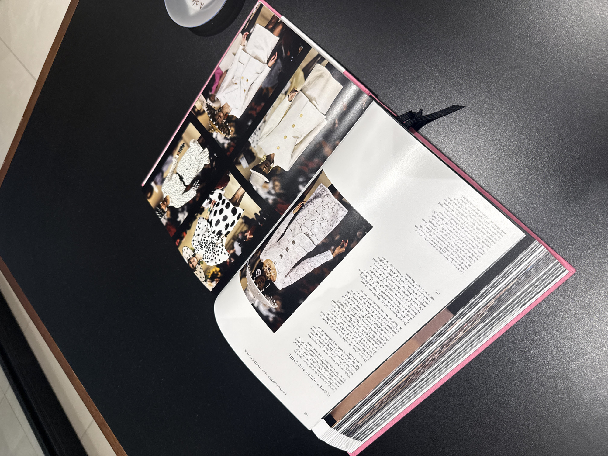The image shows an open book with a glossy black cover, resting on a dark surface. The book is angled, with its spine to the right. The pages display a collage of fashion photographs, predominantly featuring women in white outfits, some with black polka dots or intricate lacework. The garments appear to be haute couture, with elaborate designs and textures. One photograph shows a woman in a white suit with gold buttons, and another features a detailed white lace dress. The text on the right page of the book discusses a Spring/Summer 1992 haute couture collection by Yves Saint Laurent, mentioning details about the designs, fabrics, and the thirtieth anniversary of the house. The page number "454" is visible at the bottom. A black ribbon bookmark is tucked into the pages. The scene is well-lit, suggesting indoor lighting, possibly in a studio or a well-lit room, with no indication of time of day or weather. The setting appears to be a professional or personal space where one might examine a fashion book. No specific emotions are conveyed, other than a focus on the presentation of fashion.