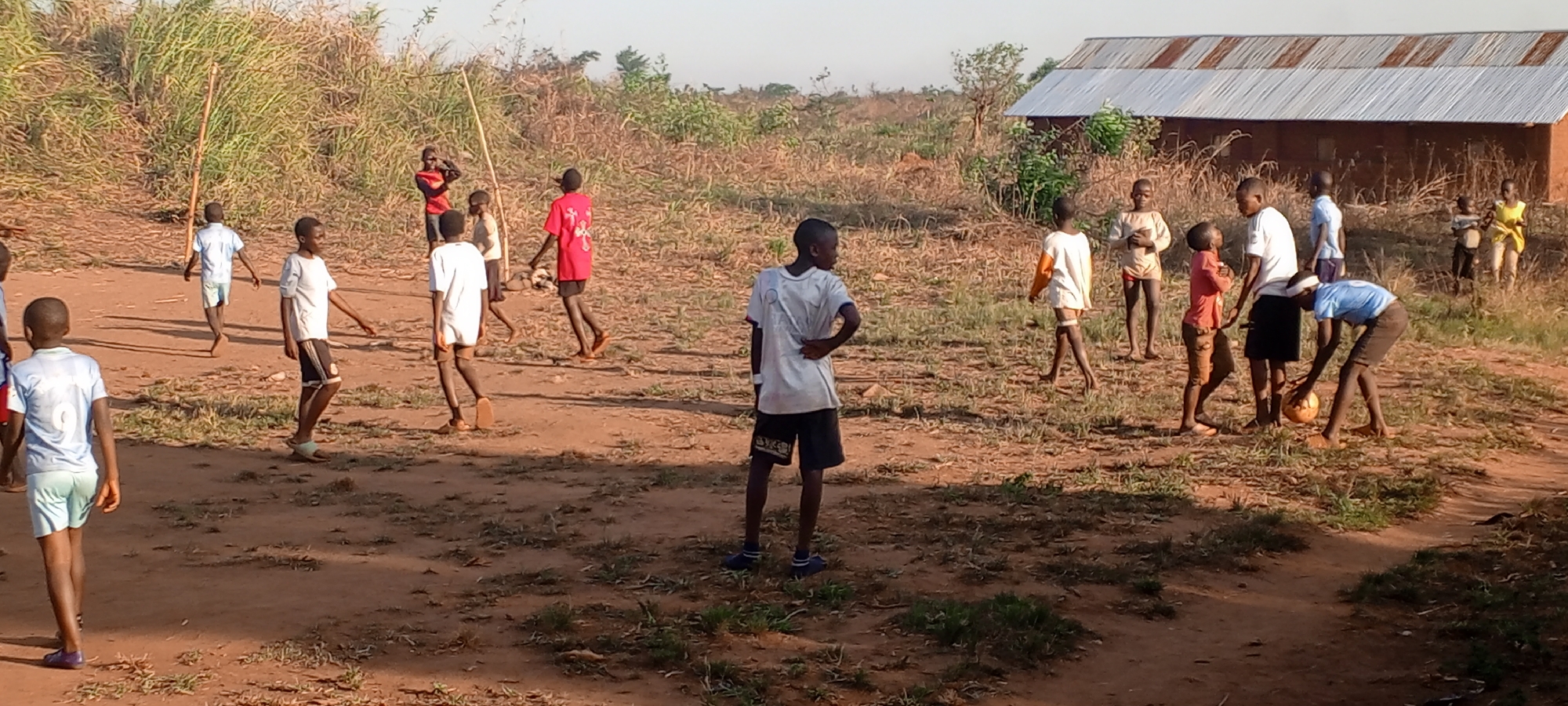 In Ngalula, Democratic Republic of the Congo, a group of children play soccer on a dirt field. The field is uneven and dotted with sparse vegetation. In the background is a small, simple building with a corrugated metal roof. The children wear a mix of clothing, mostly t-shirts and shorts. Some are barefoot while others wear sandals or shoes. Some interact with a soccer ball, while others stand and watch. A wooden pole stands upright in the distance. The scene appears to be set in a rural area, with natural vegetation surrounding the cleared field. The light suggests it is late afternoon.