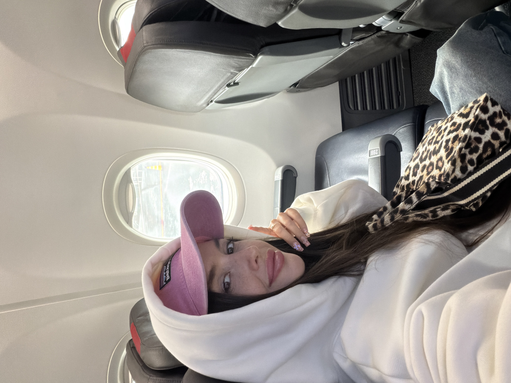 A young woman is prominently featured in a side-lying pose within an airplane cabin. She has long, dark hair, light eyes, and freckles, looking directly at the viewer with a relaxed, slightly smiling expression. Her lips are slightly parted. She wears a light-colored (likely white) hooded sweatshirt with the hood pulled up around her head, and a pink baseball cap is visible underneath the hood. Inside the cap, the brand "Patagonia" and its mountain logo are visible on a white label. Her fingernails are manicured with a detailed design.
The setting is an aircraft interior, characterized by distinct porthole windows, grey upholstered seats, and overhead panels. Through the windows, bright daylight is visible. Aircraft seats, headrests, and tray tables are apparent. Next to the woman, a leopard print bag with a black and white striped strap rests on a seat. On a seat armrest, the alphanumeric "28B" is clearly visible, indicating the seat number. The image suggests the woman is traveling and relaxing during a flight, possibly taking a selfie or being photographed. The bright lighting indicates it is daytime. The provided context indicates the location is Новое Мартьяново, Russia.