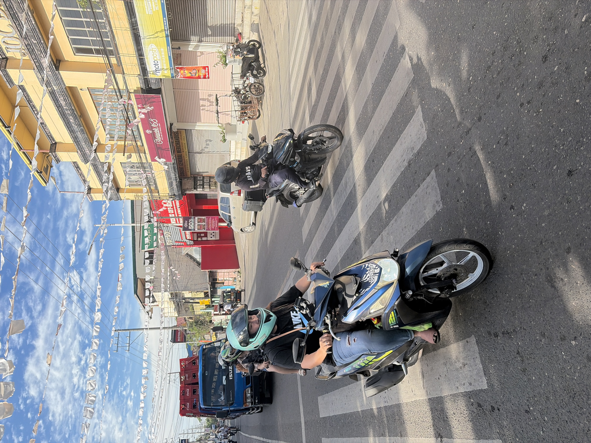 This is a photograph of a street scene in Dagupan, Philippines, likely captured during daylight given the strong shadows. In the foreground, a woman wearing a teal helmet and a black t-shirt with "NIKE" printed on it is riding a blue and black motorcycle. She is wearing ripped blue jeans and pink sandals. Her motorcycle is positioned on a pedestrian crossing. Behind her, another motorcycle is visible, ridden by a person wearing a black helmet and a black t-shirt. This motorcycle has a black storage box attached to the rear.
Further in the background, a blue truck is visible, loaded with red Coca-Cola crates. The street is lined with buildings, including a prominent yellow building with multiple windows and awnings, and other businesses with various signs, one of which reads "MOTOPOSH". White decorative flags or pennants are strung across the street and between buildings. The sky is blue with scattered clouds. The overall impression is a busy urban street with active traffic.