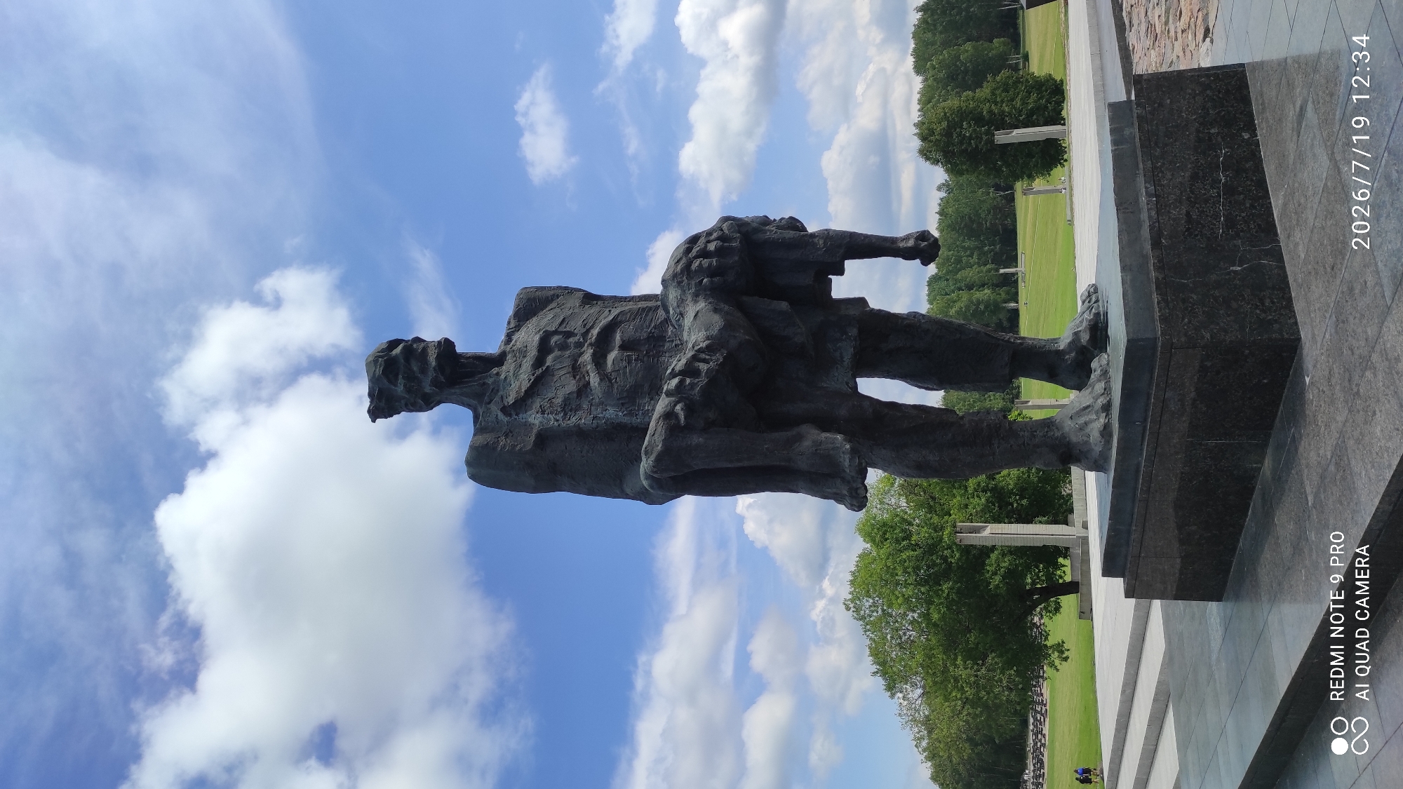 A monumental bronze sculpture depicts a gaunt, emaciated figure carrying a smaller, lifeless figure in its arms. The sculpture stands on a dark granite base, set within a large, open memorial complex under a bright blue sky with scattered white clouds. In the background, a manicured green lawn is dotted with rows of small, dark markers or tombstones, and several tall, slender stone pillars are visible. A line of trees forms the distant backdrop. The sculpture appears to represent suffering and loss, possibly related to a historical tragedy. The overall scene is somber and commemorative. The location is the Khatyn Memorial Complex, near Каменскі сельскі Савет, Belarus.