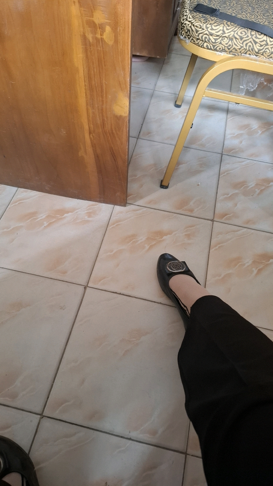 The image depicts a close-up view of a person's legs and feet on a tiled floor. The person is wearing black pants and black flat shoes with a silver buckle. A portion of a wooden table or cabinet is visible on the left side of the frame, and the leg of a yellow chair is visible on the right. The floor tiles are light-colored with a marble-like pattern in shades of pink and beige. The scene appears to be indoors, possibly in a home or office setting. There is no indication of the specific location within Bandar Lampung, Indonesia.