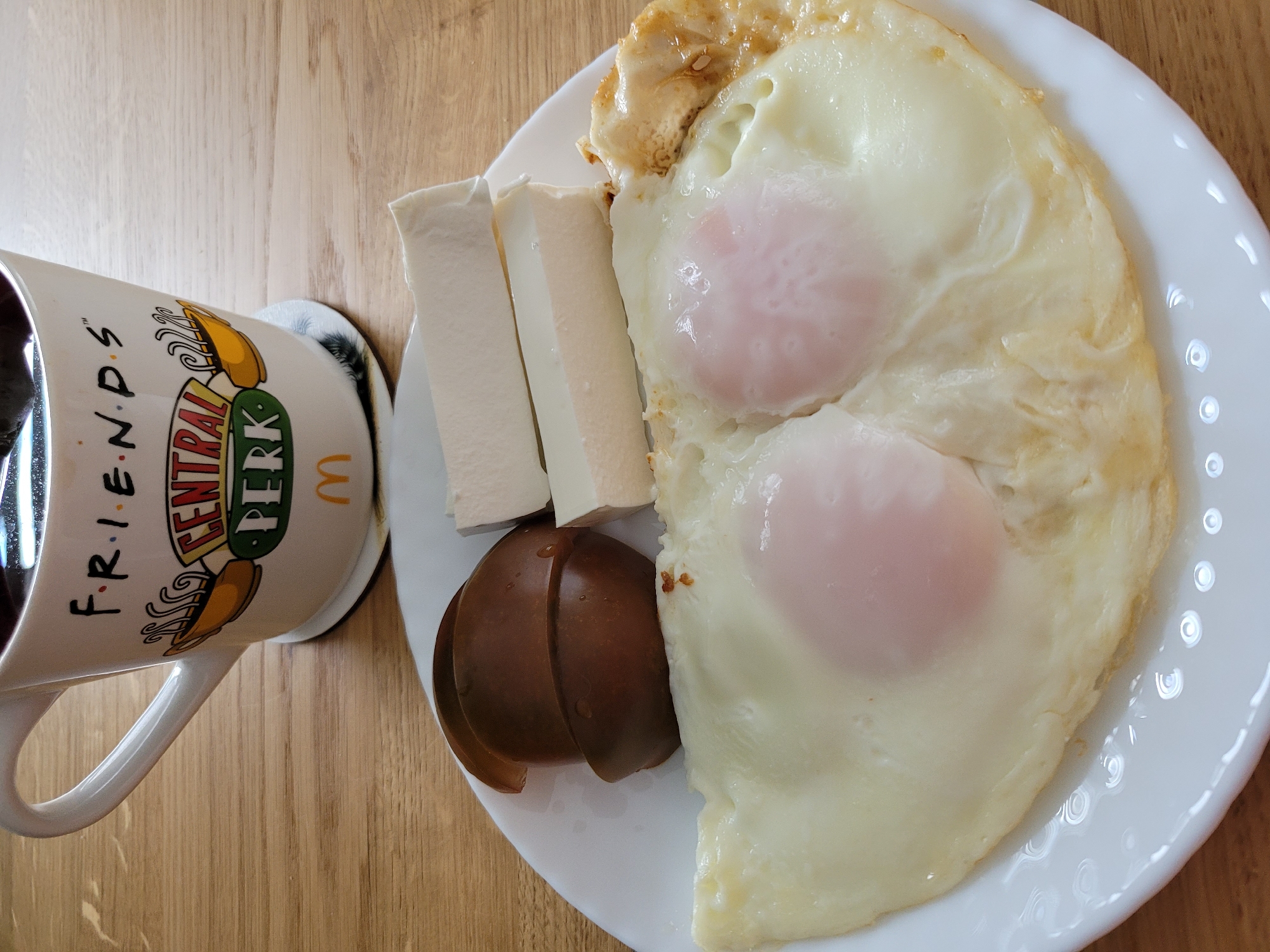 A plate of breakfast, including two fried eggs, sliced tomatoes, and cheese, sits on a wooden table next to a mug that says "Friends" and "Central Perk." The scene is set in Kyiv, Ukraine.