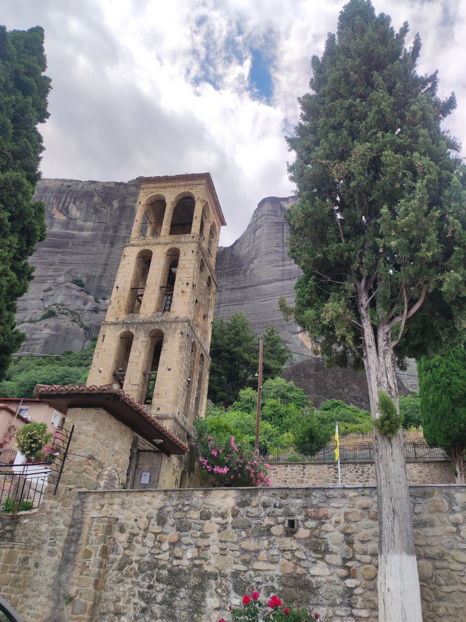 This image captures a serene scene at Meteora, Greece, showcasing a medieval bell tower rising majestically against a backdrop of imposing rock formations. The bell tower, constructed from weathered stone, features multiple arched openings and a distinct layered design. To its left, a small stone building with a terracotta tiled roof is partially visible, adorned with pink flowering bushes. A sturdy stone wall with varying sized stones forms the foreground, leading the eye towards the historical structures.
The surrounding environment is lush with greenery, including tall, conical cypress trees flanking the bell tower and a variety of other trees and bushes cascading down the slopes. The massive, striated rock formations of Meteora dominate the background, their sheer faces hinting at the area's geological significance.
The sky is overcast with scattered clouds, suggesting a temperate, possibly early afternoon, atmosphere. There is no visible activity of people. The overall emotion evoked by the image is one of timelessness and natural grandeur, harmoniously blended with human architectural endeavors. A small yellow flag flies from a pole in the middle ground, adding a subtle touch of color to the scene. No other visible text is present in the image.