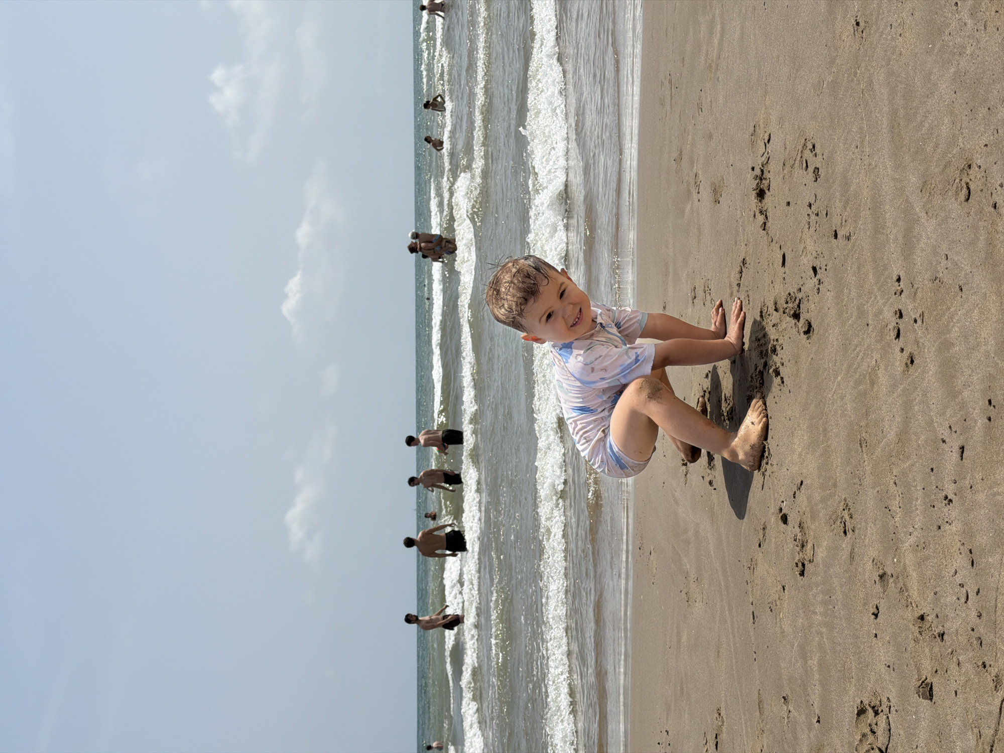 A young boy with sandy brown hair and a light blue and pink striped rash guard is crouched on a sandy beach, facing the camera and smiling. His knees are bent, and his hands are on the sand in front of him. Several people are visible in the ocean behind him, wading and swimming in the waves. The sky is a light blue with a few scattered white clouds. The scene appears to be a sunny day at the beach.