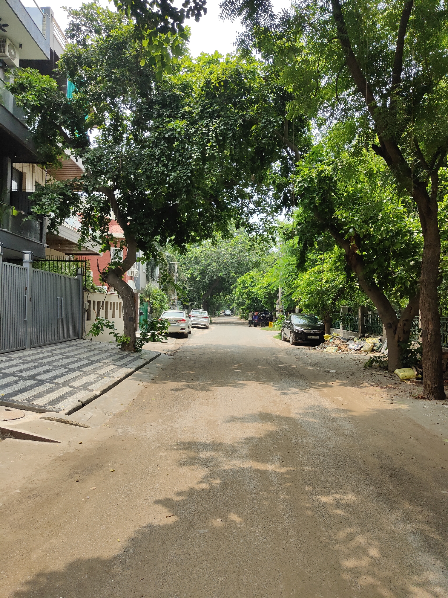 The image depicts a residential street in Gurgaon, India, on a clear day. The street is paved and runs between houses and trees. On the left side of the street, a modern house with a gray facade and large windows is visible. A gray metal gate and a patterned walkway are in front of the house. A large tree with dense green foliage partially obscures the view of the house and overhangs the sidewalk. On the right side of the street, several cars are parked, including a black sedan and two white sedans. Piles of construction debris and yellow plastic bags are also present along the right side of the road. The street is lined with mature trees casting shadows on the pavement. In the background, more houses and trees create a verdant urban landscape. No people are clearly visible in the image, though a utility cart is parked further down the street on the left.