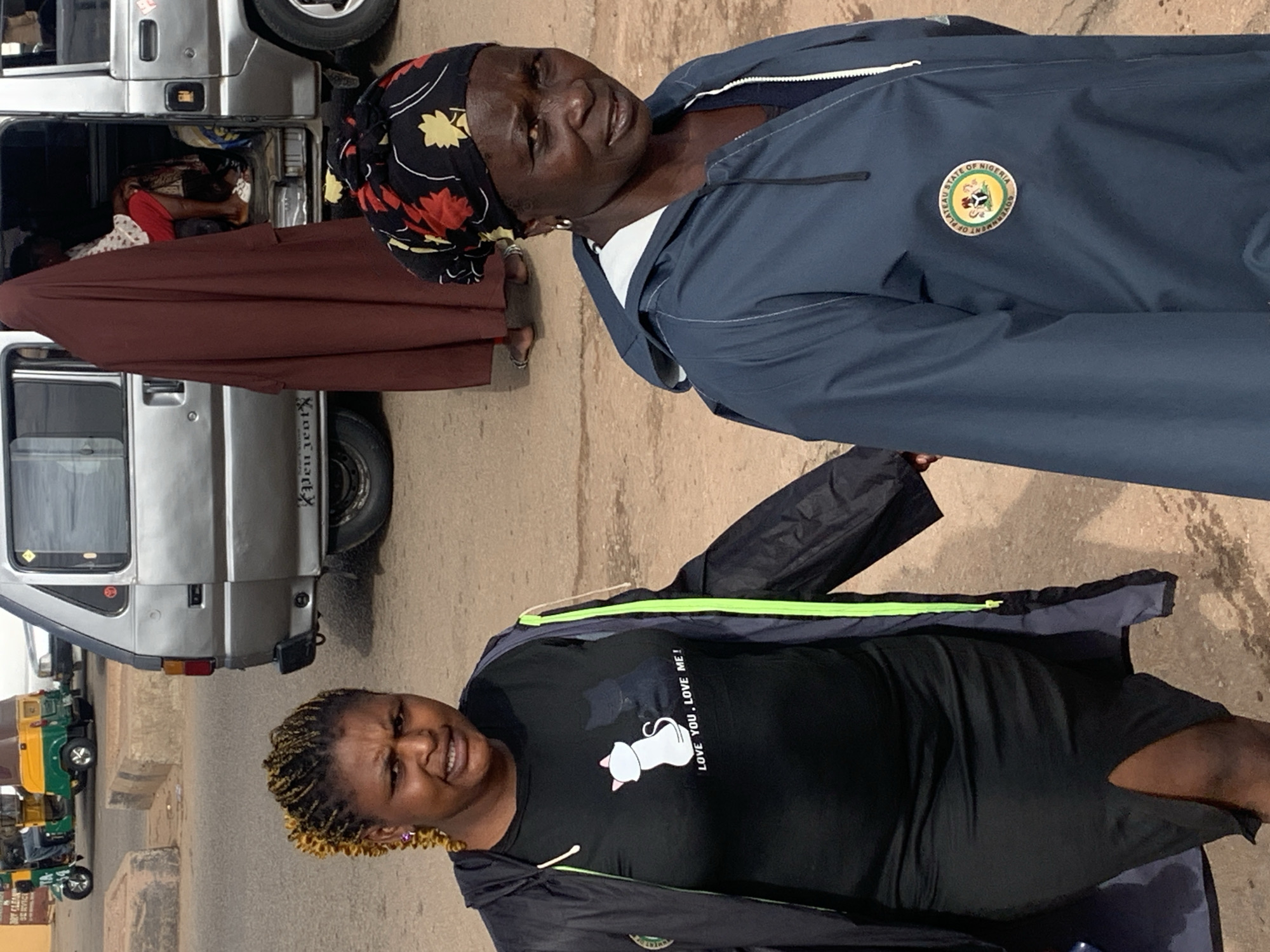 In the city of Gura Top, Nigeria, two women are walking on a dusty, outdoor street. The woman on the left, with blonde braided hair and wearing a black t-shirt with a cat graphic and a black jacket with neon green accents, is looking towards the camera with an open mouth. The woman on the right, wearing a dark blue jacket and a colorful headscarf, is looking towards the camera with a neutral expression. In the background, a silver van and a colorful auto-rickshaw are parked. A person in a dark brown garment is standing near the van. The scene appears to be a street market or a busy thoroughfare.