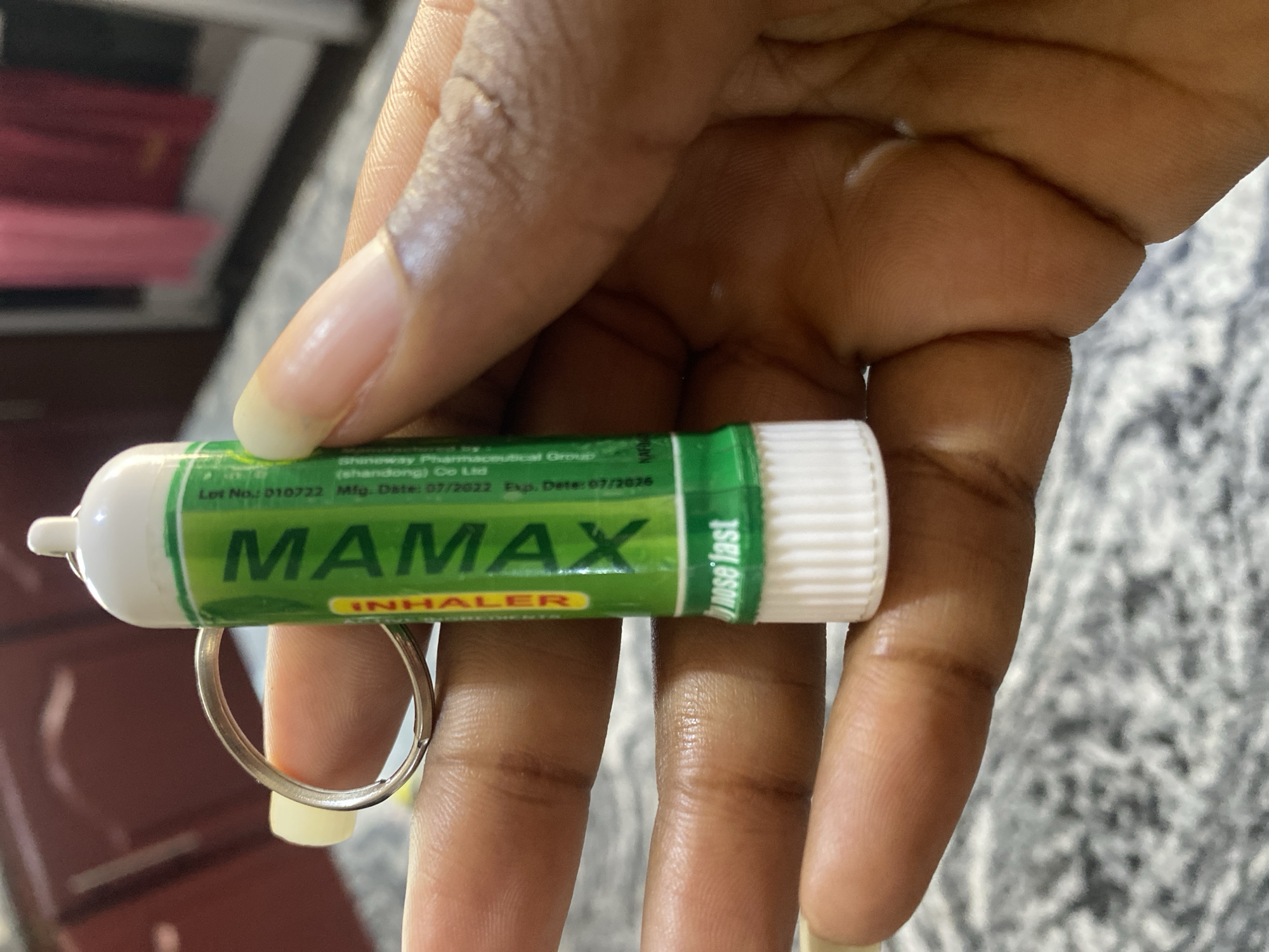 A person's hand, with dark skin and short, light-colored fingernails, holds a green and white nasal inhaler. The inhaler has "MAMAX" printed in large, bold, white letters. Below that, the word "INHALER" is printed vertically in yellow. Text in smaller white print on the green section reads "Lot No: 010722 Mfg. Date: 07/2022 Exp. Date: 07/2026" and "Graceway Pharmaceutical Group Co Ltd". A white cap with a textured surface sits at the bottom of the inhaler. A silver keyring is attached to the top of the inhaler. The background is blurred, but appears to be indoors, with hints of a dark red cabinet and a pink fabric hanging. The scene is located in Bar Jirgi Summa, Nigeria.