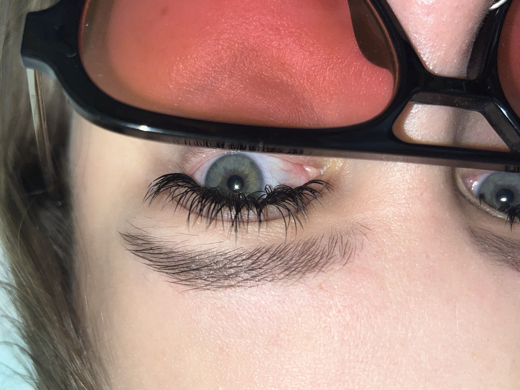 A close-up, eye-level shot captures the upper face of a person, focusing intently on their eyes and a pair of distinctive glasses. The individual is wearing thick, black-framed eyeglasses with large, reddish-orange tinted lenses that dominate the upper portion of the frame, obscuring the forehead and much of the upper face. The inside surface of the lenses shows a textured reflection, possibly of skin or an indoor light source.
The left eye is prominently featured, revealing a striking light green or hazel iris with a clearly defined dark pupil. The white of the eye shows minor, faint red capillaries. Extremely long, dark, and voluminous eyelashes, indicative of lash extensions or false lashes, frame the upper and lower eyelids. Beneath the eye, a well-groomed, natural-looking dark eyebrow with discernible individual hairs is visible. The skin around the eyes and on the visible cheek appears fair with natural pores and texture. A section of light brown hair is visible on the far left. The right eye is partially visible towards the right edge of the frame, also framed by dark lashes.
The image presents a detailed portrait, emphasizing the eyes and the unique eyewear. No specific activity is clearly depicted beyond being photographed. There is no visible text within the image. The context provided indicates the location is Krasnodar, Russia.