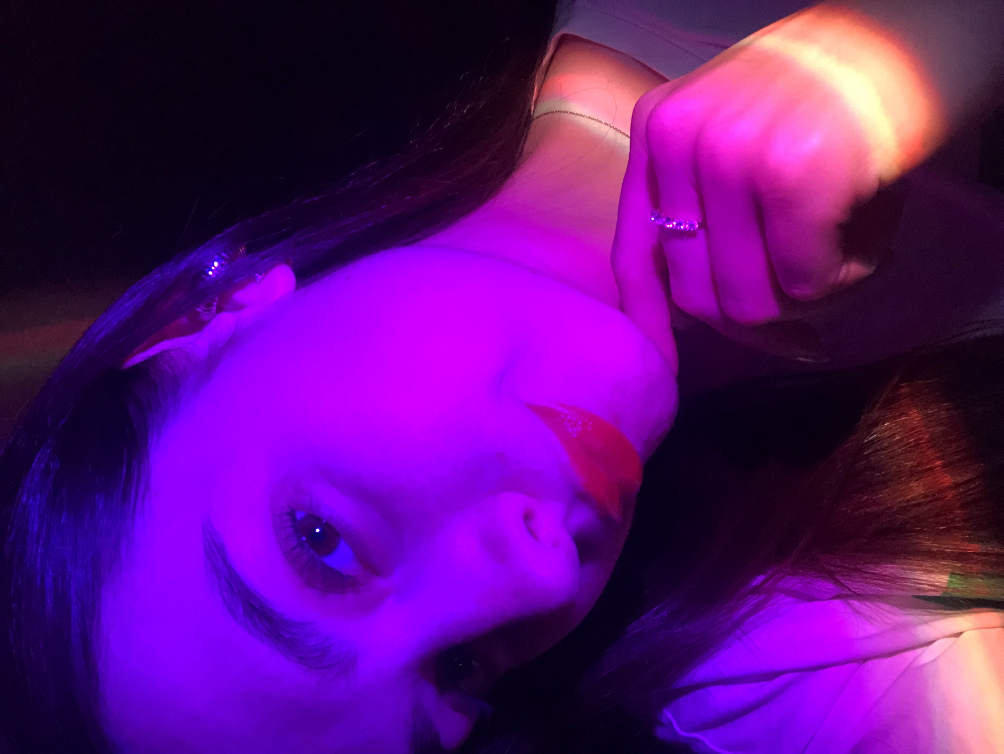 A close-up, rotated image shows a person's face and hand under purple and orange lighting. The person has dark hair and is wearing bright red lipstick. A ring is visible on one of their fingers. The background is dark. The person is holding their neck. The image is poorly lit. No location of Noviy, Russia is verifiable.