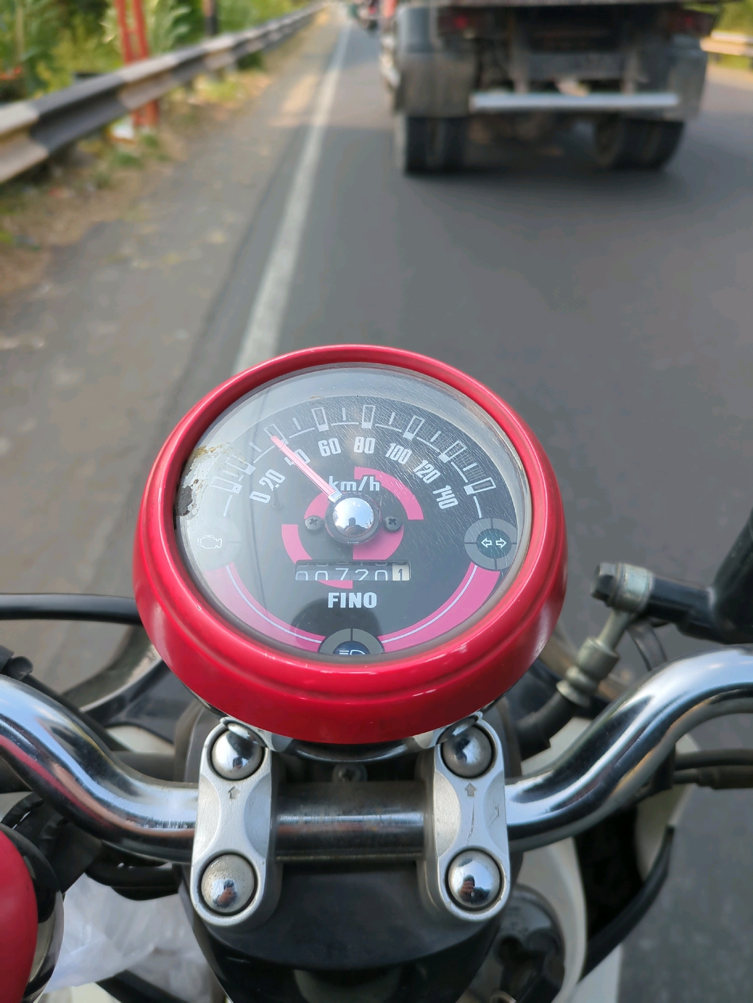 The image is a POV shot from a motorcycle on a road in Desa Ngesrep, Indonesia. The motorcycle's speedometer is prominently displayed, showing a speed of approximately 40 km/h. The motorcycle has a red bezel around the speedometer and chrome handlebars. In the background, a large truck is visible driving on the same road. The road appears to be paved and has a white line marking the lane. A guardrail runs along the side of the road, with greenery visible beyond it. The overall lighting suggests it is daytime.
