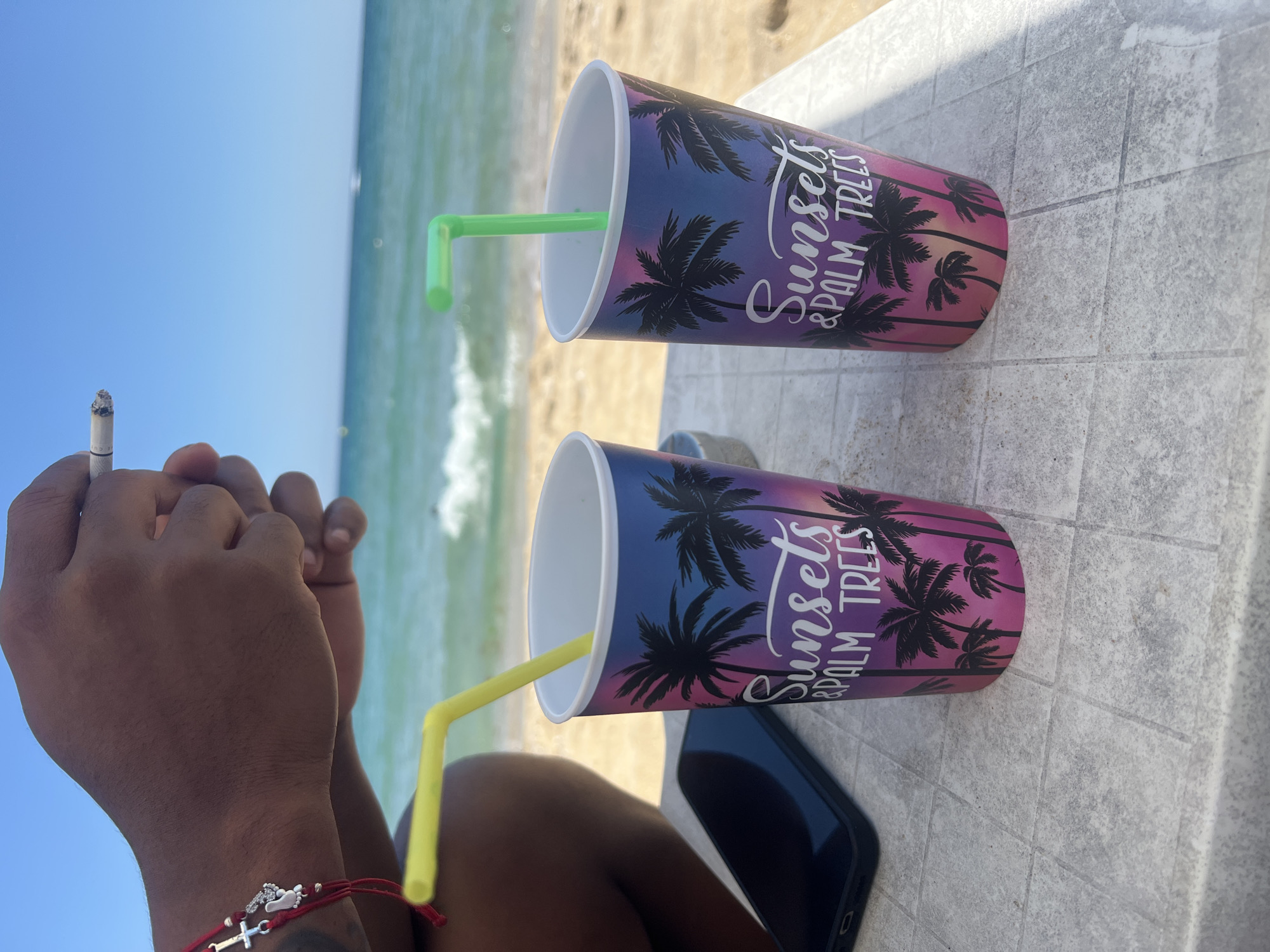 Two paper cups with the design "Sunsets & Palm Trees" stand on a tiled table near the sea. A yellow straw is in the left cup, and a green straw is in the right cup. The sea with waves is visible in the background, along with a sandy beach. A person's hands are in the upper part of the image, one holding a lit cigarette. A smartphone lies on the table to the left of the cups. The scene is set on a sunny day.