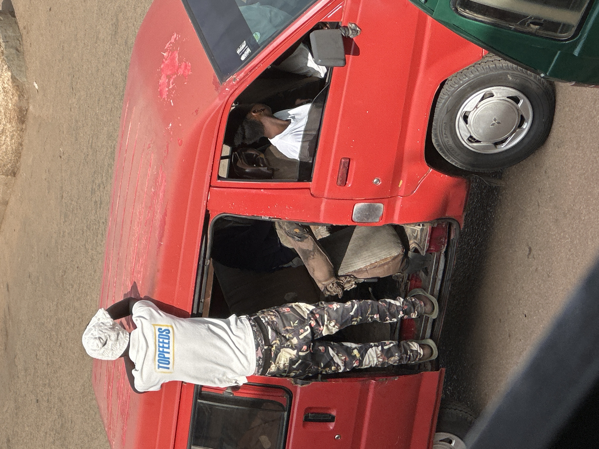 In Bukuru, Nigeria, a man wearing a white beanie and a white t-shirt with "TOPFEEDS" printed on it in blue and yellow stands with his back to the camera, leaning over the roof of a bright red van. He is positioned near the open driver's side door, with his left leg inside the van. His pants are dark with a camouflage pattern featuring floral and bird motifs. Inside the van, another person wearing a white t-shirt is visible through the driver's side window, looking to the right. The van's interior appears to be filled with objects, including a large, brown, possibly fabric-covered item that extends from the back. The scene takes place on a paved road, with a dusty, light-colored surface visible in the background. A portion of a green vehicle is visible in the lower right corner of the image.