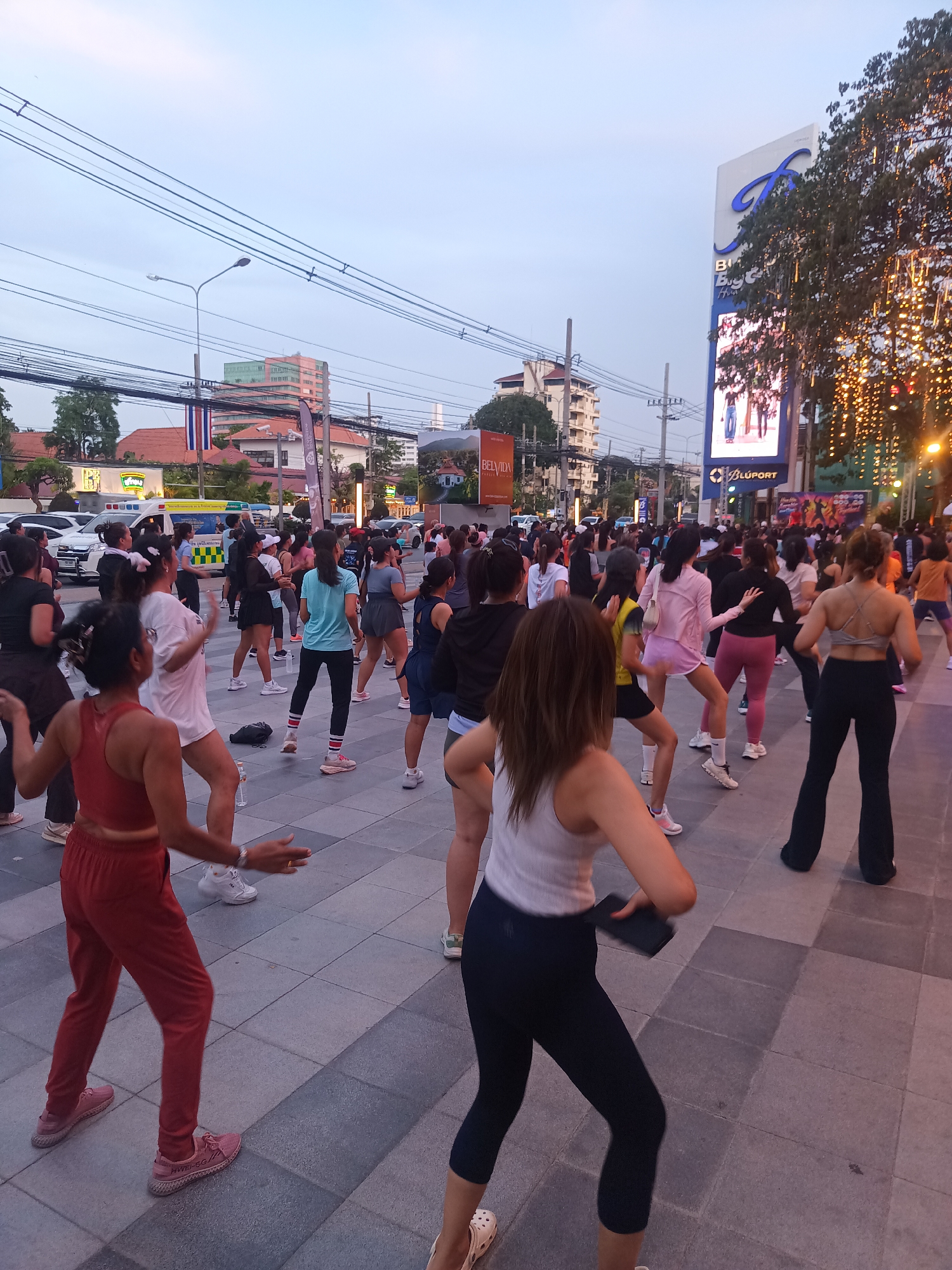 An outdoor aerobics class is taking place in Hua Hin City Municipality, Thailand, on a plaza paved with square tiles. The participants, mostly women, are dressed in sportswear and sneakers, and are engaged in synchronized movements. They are facing away from the viewer towards a brightly lit commercial area. A large digital billboard displays images of people. Behind the participants are cars, a Thai flag, and buildings, suggesting an urban setting. Overhead, there are electrical wires. The sky is a pale dusk color.