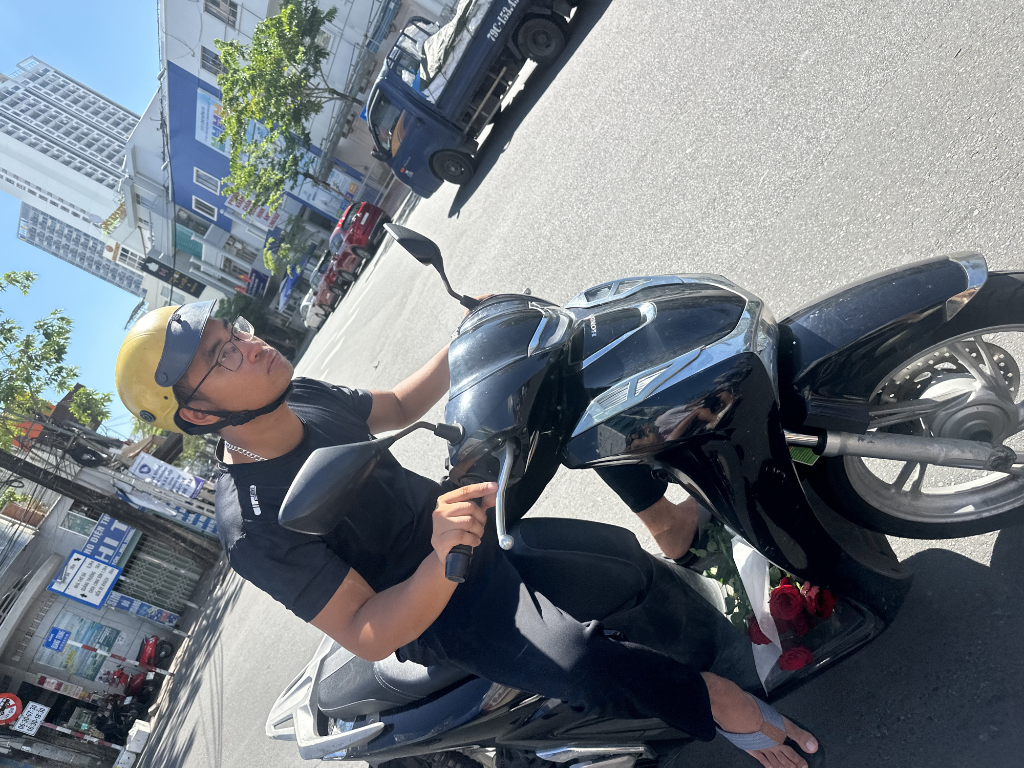 In Nha Trang Ward, Vietnam, a man wearing a yellow helmet and glasses rides a black scooter on a sunny day. He is dressed in a black t-shirt and black pants, with his feet clad in gray flip-flops. In front of him, on the scooter, are a bouquet of red roses wrapped in white paper. The scooter is on a paved street, with buildings, trees, and other vehicles visible in the background, including a blue truck with license plate "79C-153.45". The overall scene depicts a mundane urban street view with a personal touch of the flowers.