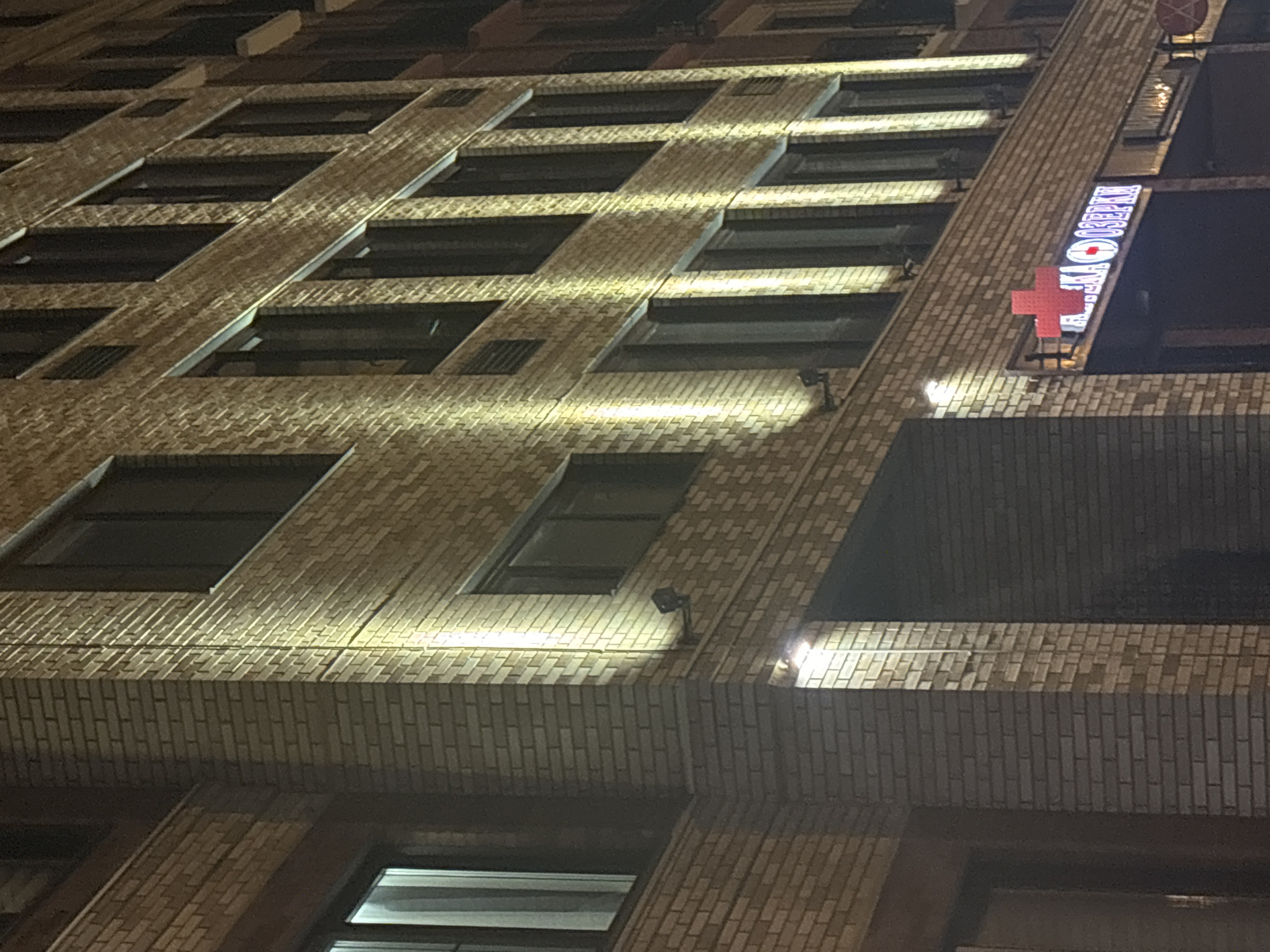 The image shows the exterior of a brick building at night, illuminated by yellow lights. The building has multiple stories with numerous dark windows. On the lower level, a red cross symbol is visible above a sign with Cyrillic lettering. The sign appears to be for a pharmacy. The overall scene suggests an urban environment. Based on the Cyrillic lettering on the sign, the location is likely in Russia, possibly in the city of Novosibirsk.