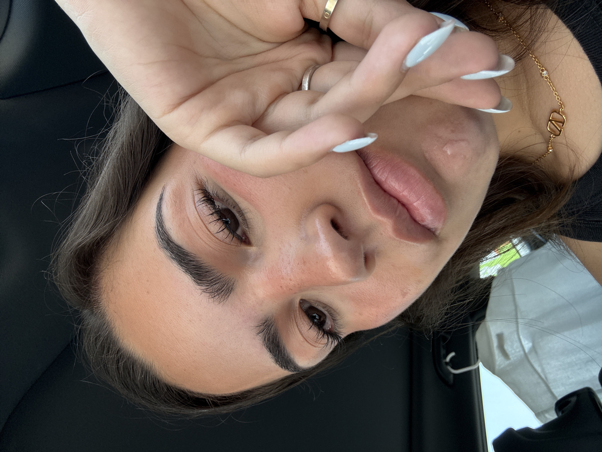 A woman is in a car. She has brown hair, fair skin, and manicured fingernails with a light blue polish. She is wearing a gold necklace and rings. Her hand is partially covering her face. A disposable face mask hangs from the car's interior. The location cannot be determined.