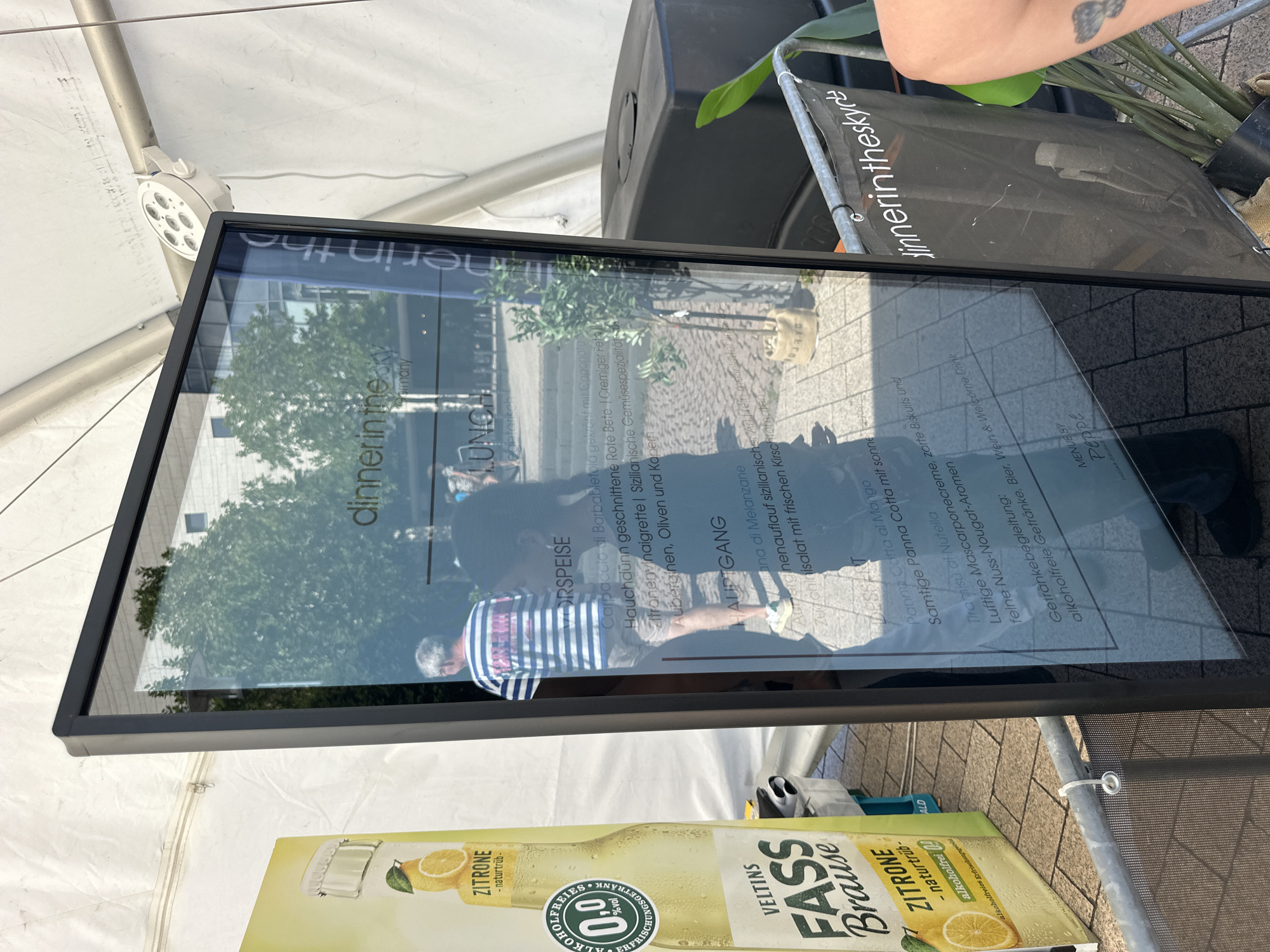 The image shows a digital menu board set up outdoors, likely at an event in Cologne, Germany, given the visible text. The menu board displays "dinner in the sky Germany" at the top, and lists "Vorspeise," "Hauptgang," and "Dessert" with Italian and German descriptions of the dishes. To the left of the menu board is a large bottle of "VELTINS FASS BRAUSE Zitrone" with an "ALKOHOLFREIES 0,0% vol" label. In the background, reflections on the menu board show trees and buildings, suggesting an urban outdoor setting. There are people visible, including a man in a striped shirt and another person whose face is obscured by reflection. A black mesh banner with "dinnerinthesky" written on it is attached to the front of the menu board. The scene is illuminated by natural daylight, and a white spotlight fixture is visible attached to a tent structure overhead.