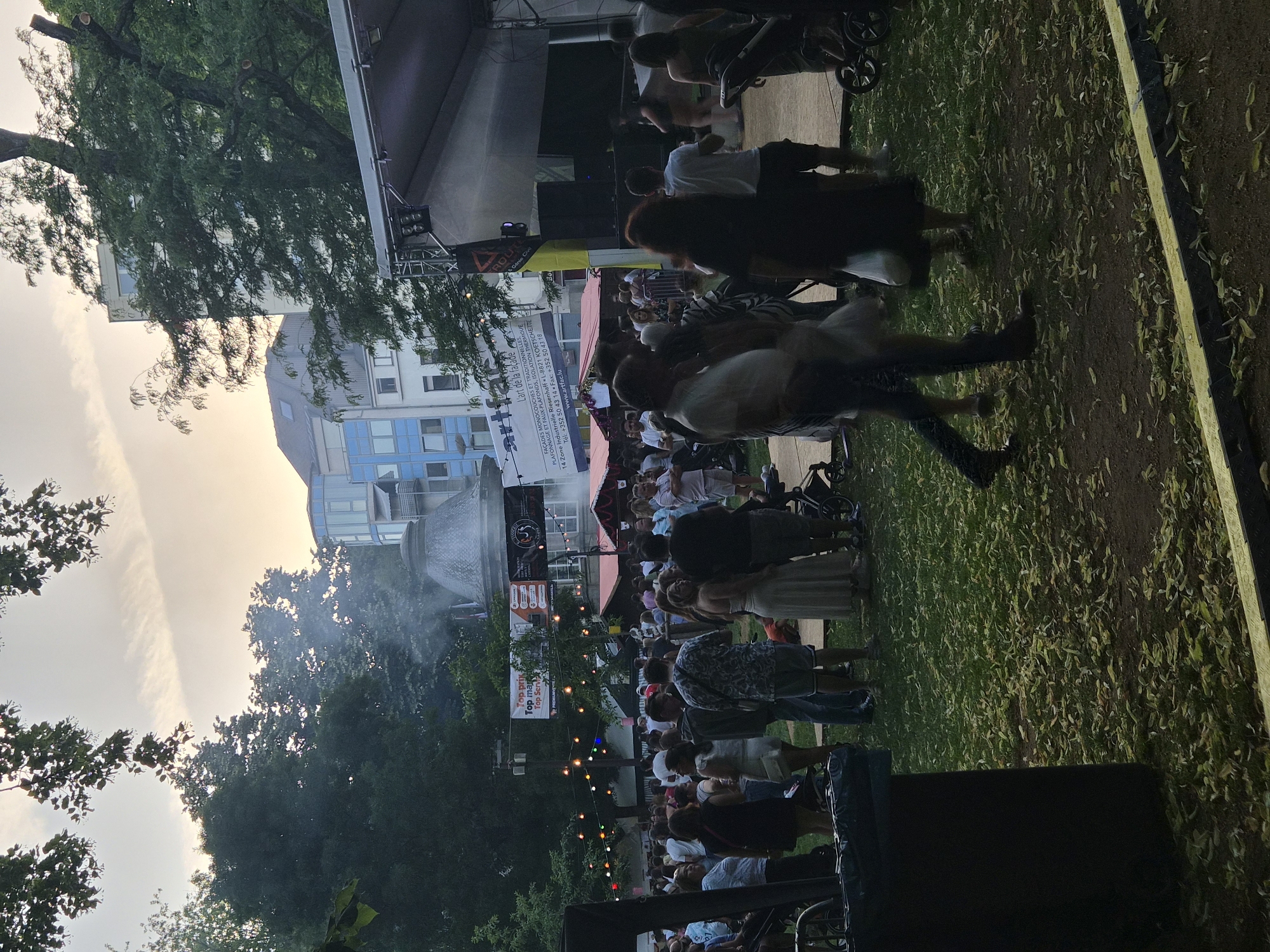 The image captures an outdoor event, likely a festival or market, set in a park-like environment within the city of Differdange, Luxembourg. The scene is populated by a diverse group of people, appearing to be a mix of locals and visitors, engaged in various activities. In the foreground, several individuals are standing, some in conversation, while others are observing the surroundings. Children are also present, with some being pushed in strollers.
The environment features a grassy area with scattered leaves, suggesting an autumnal season or a park with mature trees. In the background, a stage structure with lighting equipment is visible, indicating a performance or event is taking place. Numerous stalls or temporary structures are set up, possibly for food vendors or merchandise. String lights are strung across the area, adding a festive ambiance. Beyond the event space, modern buildings with glass facades and balconies are visible, characteristic of an urban setting. Wisps of smoke or mist are rising from a dome-shaped structure in the mid-ground, which could be a fountain or part of the event's attractions. The sky above is partly cloudy, with the soft light suggesting either early morning or late afternoon. The overall atmosphere appears lively and social, with people enjoying the outdoor gathering.