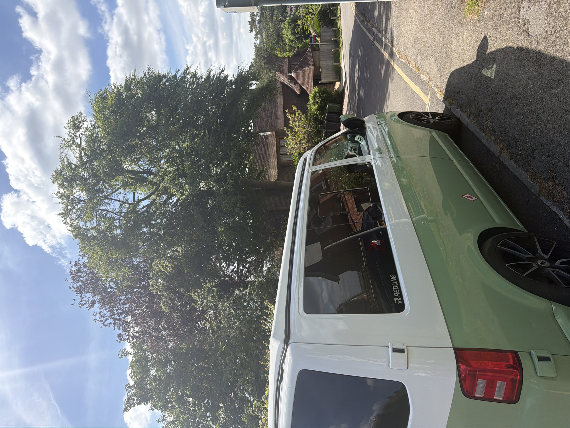 The image captures a street scene in Guildford, United Kingdom, featuring a two-tone light green and white Volkswagen Transporter parked alongside a road. The road is marked with a yellow line along the curb. The vehicle's side is prominently displayed, including details like the "REDLINE" logo on a window. A large tree dominates the left side of the frame, partially obscuring a house in the background. The sky is visible above the tree, featuring scattered clouds and bright sunshine. A shadow is cast on the road near the vehicle, and portions of a building can be observed behind the tree and the houses.