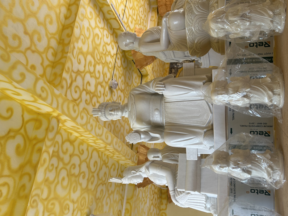 An interior scene in Đăk Hring, Vietnam, displays multiple white statues beneath a bright yellow ceiling adorned with repeating golden cloud patterns and decorated beams. In the mid-ground, a prominent, large white statue of a male figure is centrally seated, wearing an elaborate headdress and traditional robes, holding a circular emblem with a symbolic design in his raised right hand. To his left, a smaller white female statue, also in traditional attire and a headdress, sits in profile, facing right. Behind her, a partially visible, bald, seated figure is present. To the right of the central male statue, another smaller, bald, white statue resembling a Buddha or monk sits in profile, facing left, on a lotus-shaped base. In the foreground, several smaller, identical white statues of kneeling or standing figures, possibly young monks, are individually wrapped in clear plastic bags. These foreground statues rest on stacks of rectangular panels marked "Reto ALUMINUM COMPOSITE PANEL," indicating temporary storage or display. The overall lighting is bright and even.