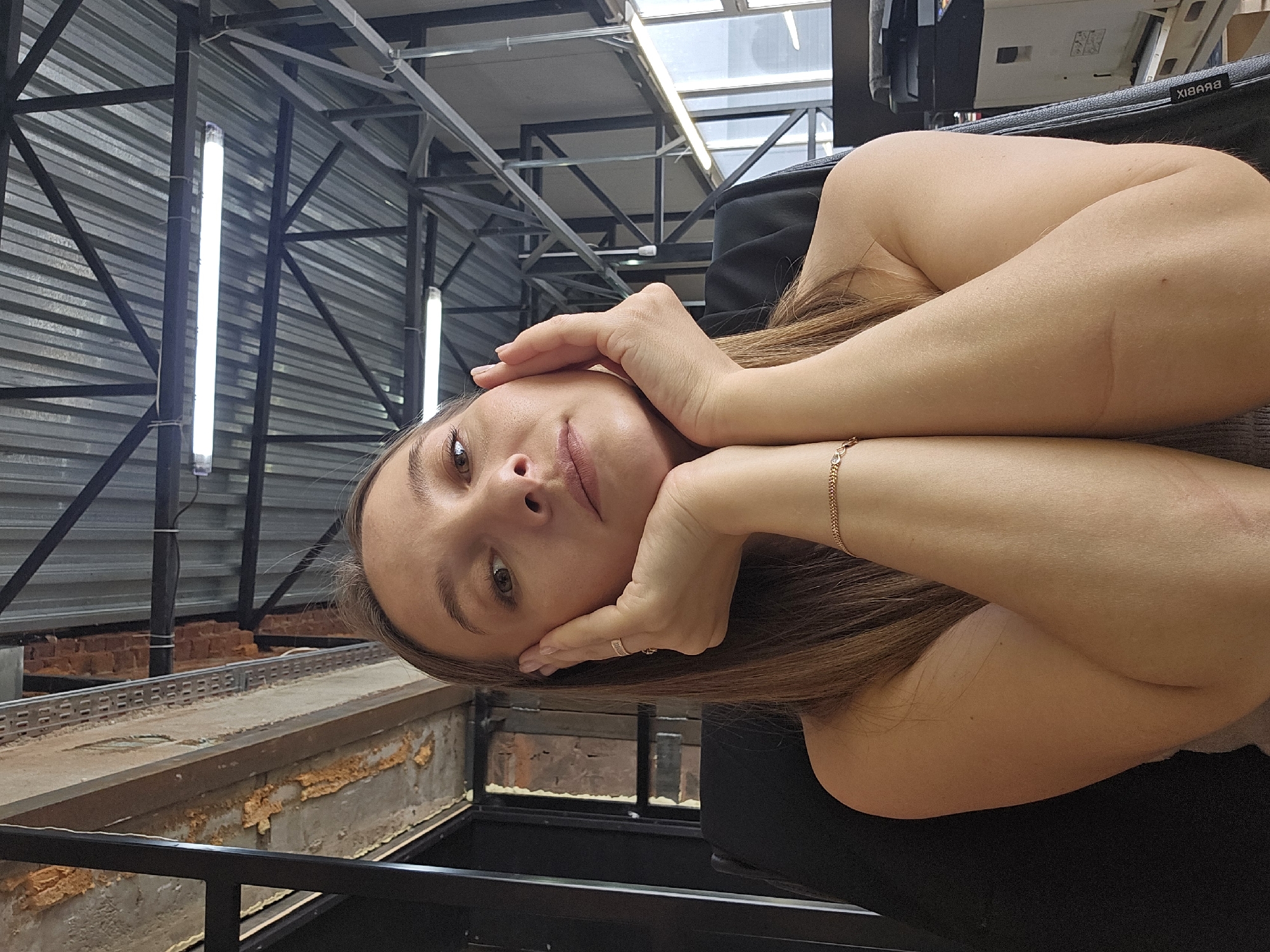 An adult female is depicted seated indoors in an industrial-style commercial or office building located in Chaikovsky, Russia. The subject faces the camera directly, exhibiting a neutral to slightly serious expression. Her hands are raised, symmetrically cupping her cheeks with thumbs resting near her chin. She has brown, medium-length hair and fair skin. Her attire includes a light-colored, possibly sleeveless top. She wears a thin gold-colored chain bracelet on her right wrist and a ring on the fourth finger of her left hand.
The background features a high, exposed ceiling constructed from corrugated metal panels supported by dark-colored metal trusses and beams. Several illuminated fluorescent tube lights are suspended from this structure. To the upper left, an exposed concrete beam with orange spray foam insulation and reddish-brown brickwork is visible, framed by dark metal supports. To the upper right, large windows or glass panels allow natural light, with a portion of what appears to be a printer or copier visible through them in the distance. The subject is seated in a black office chair, on the side of which the brand "XIBBIX" is legible.