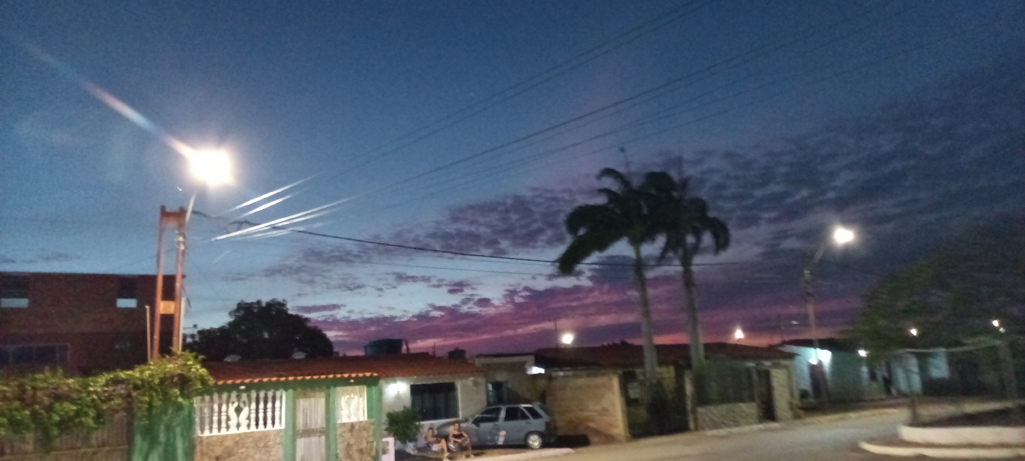 The image depicts a street scene in Unknown, Unknown, at dusk or night. The sky transitions from blue to a band of pink and purple near the horizon, partially obscured by thin, textured clouds. Utility lines stretch across the sky.
In the foreground, a street lamp illuminates the left side of the frame, casting a glow on a building with a fence covered in vegetation. A car is parked near a house with a red-tiled roof. Two palm trees are silhouetted against the colorful sky. Further down the street, additional houses and a street lamp can be seen. The street itself is dimly lit and leads away from the viewer. No people are readily visible.
