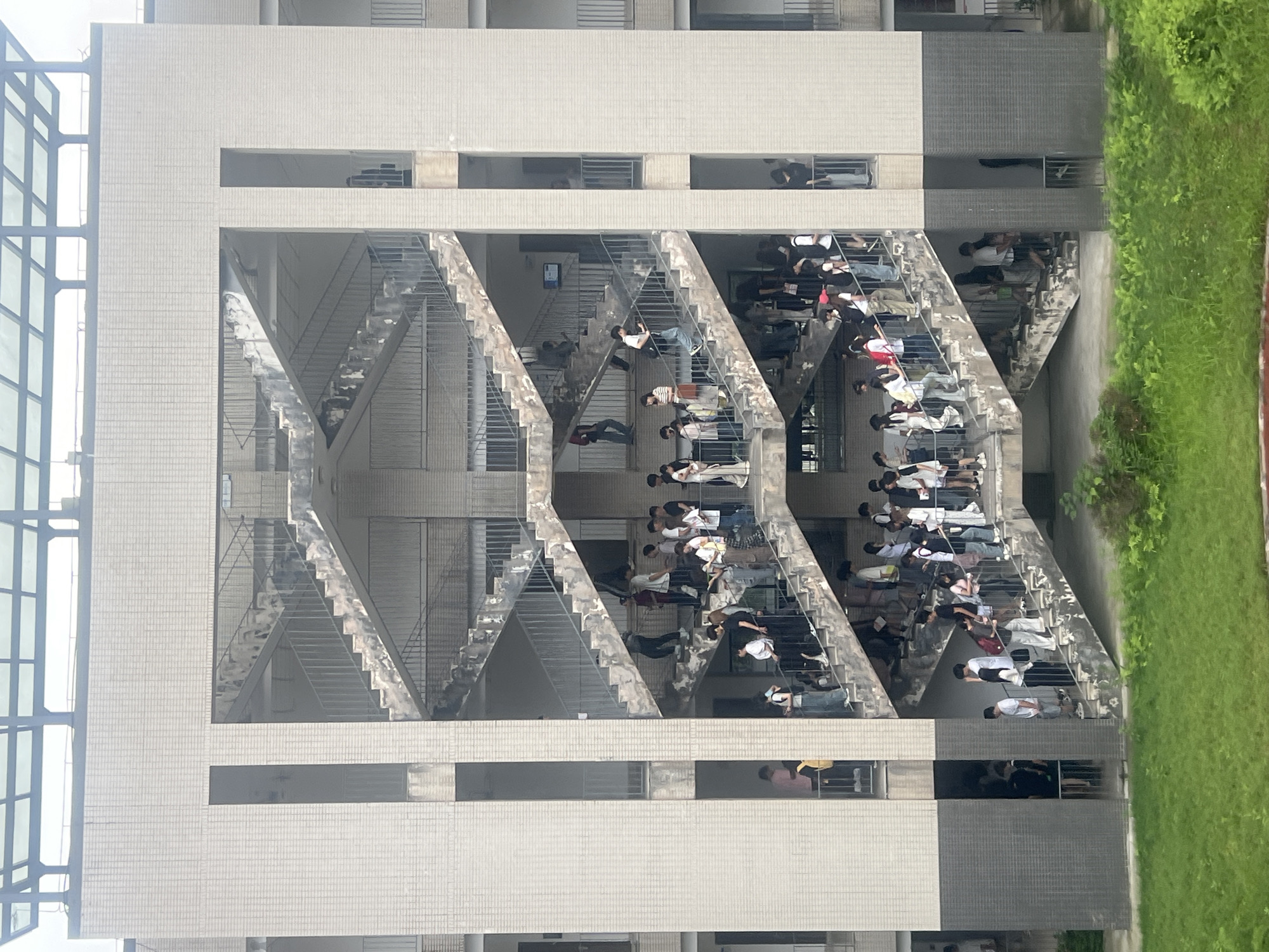 The image depicts a multi-story building with exposed staircases on the exterior. Numerous individuals are observed sitting on the staircases, some appearing to be reading or studying. The building is constructed with light-colored bricks and has square windows. To the left of the building is a glass structure. To the right is green vegetation. The context of the image, based on the location, may indicate the photo was taken in the city of Jiangjin, China.