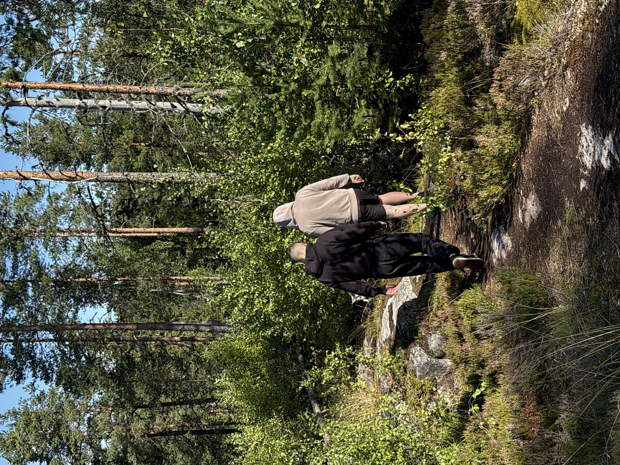 Two individuals, viewed from behind, are walking along a dirt path through a sun-dappled pine forest. The person in the foreground wears a black hooded sweatshirt and dark pants, while the person ahead is in a beige hooded sweatshirt and dark shorts. They are in a densely wooded area with tall, mature pine trees and thick undergrowth of green shrubs and heather. The ground is uneven, with rocks and patches of moss. The scene appears to be located in Kir'yavalakhti, Russia, indicated by the environment typical of the Karelian region.