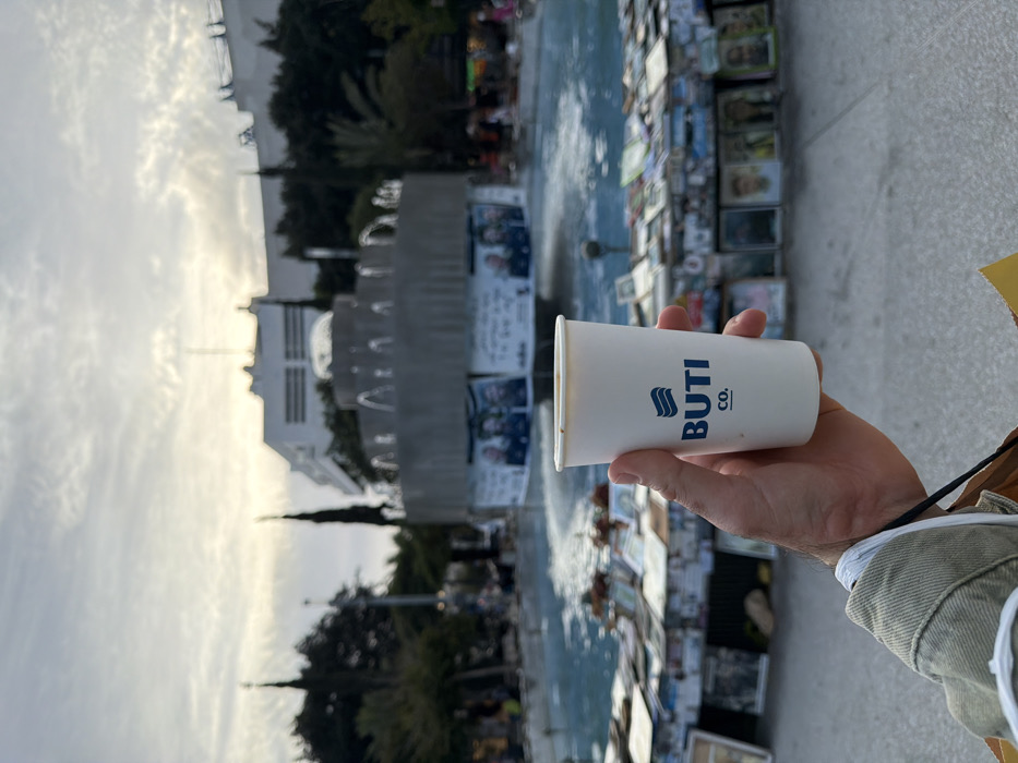A hand holds a white paper cup with “BUTI CO.” printed in blue. The background shows a tiered fountain decorated with photos and signs. Trees and a white building are visible in the distance under a cloudy sky. The scene appears to be outdoors during daylight, possibly near a memorial or protest site. The photograph is slightly rotated.