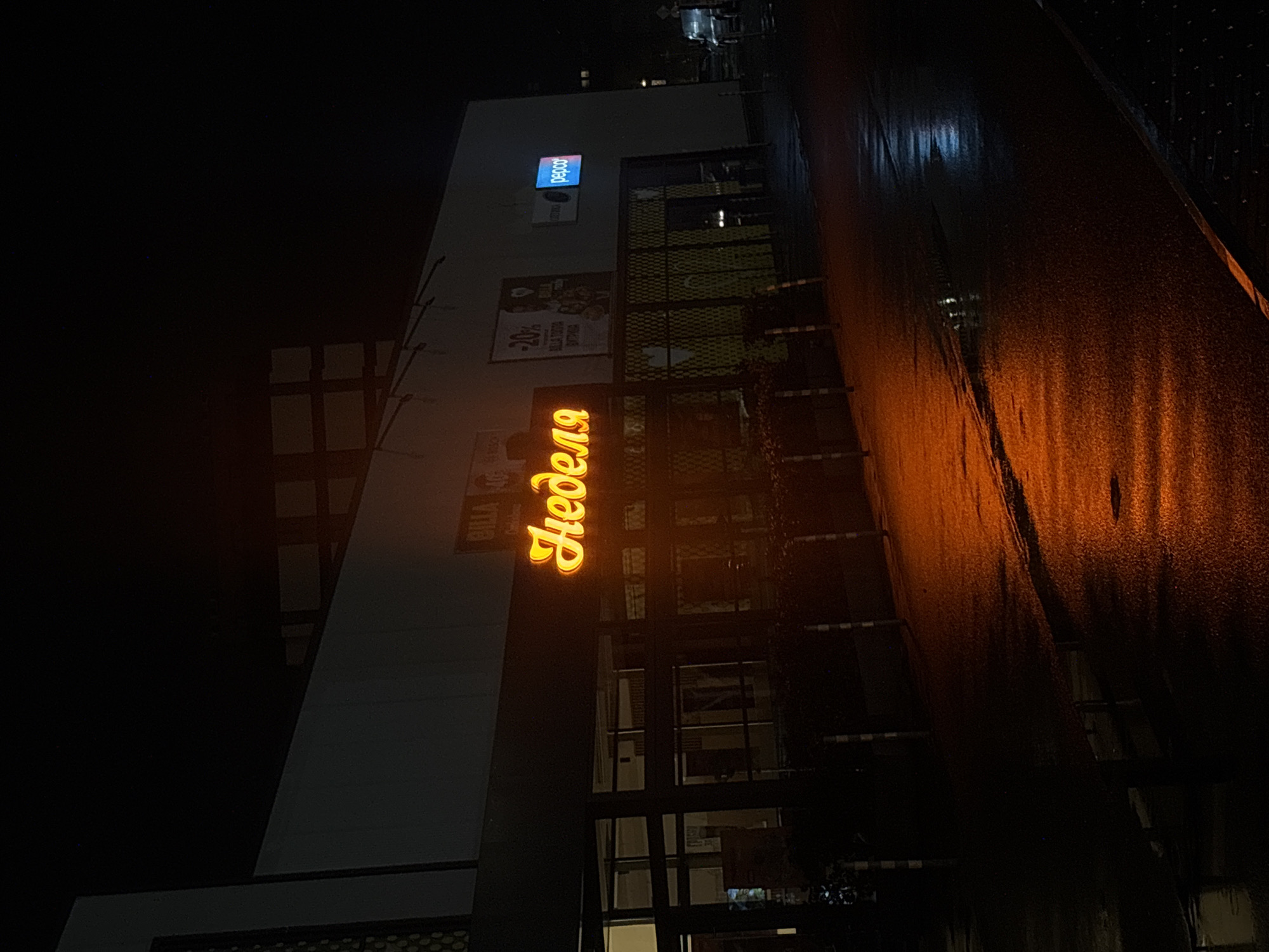 The image depicts the exterior of a building at night in Sofia, Bulgaria. Illuminated signage on the building reads "Неделя" vertically in a distinct golden-yellow color. Above this sign, there's a smaller poster with the words "BILLA" and "Bulgaria". Higher up on the building, a "pepco" sign is visible. The street surface appears wet, reflecting the ambient light. The surrounding environment is dimly lit, with adjacent buildings and structures partially visible in the darkness. The overall impression is a nighttime urban scene focusing on commercial establishments.