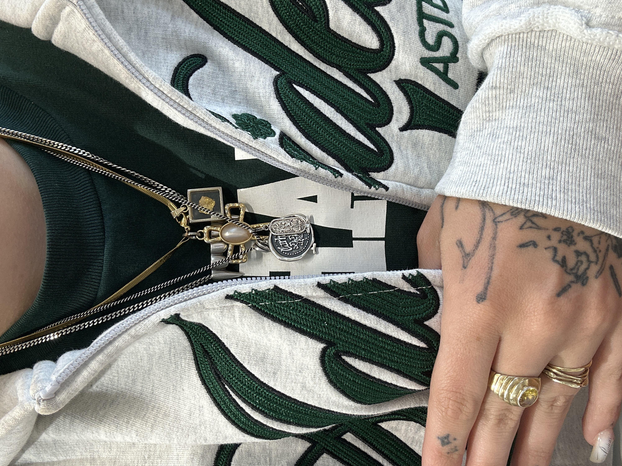 Close-up view of a person wearing a gray zipper hoodie with dark green lettering, along with multiple gold and silver necklaces. The person is also wearing a dark green top underneath the hoodie. Two gold rings are visible on the person's fingers, and there are tattoos on their hand and arm. The image does not provide sufficient information to determine the location in Paris, France.