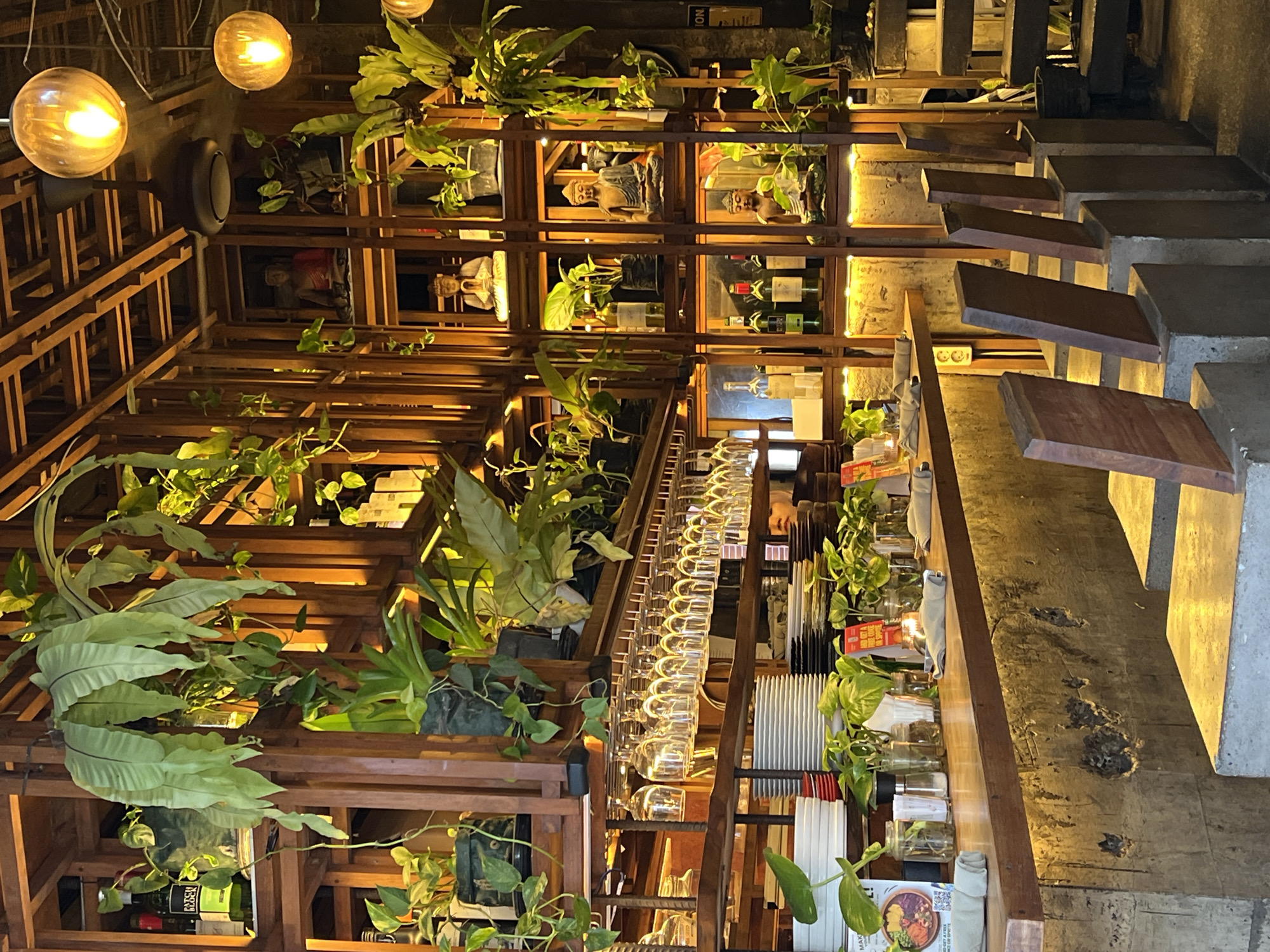 This image depicts a warm and inviting interior space, likely a restaurant or bar, characterized by an abundance of natural elements and soft lighting. The scene is dominated by a multi-tiered wooden shelving system that fills the left side of the frame, from floor to ceiling. This structure is heavily adorned with various green plants, including large-leafed ferns and vining foliage, creating a lush, organic atmosphere.
Within the wooden shelves, several Buddha statues are visible, some in seated meditative poses, contributing to a tranquil, culturally resonant ambiance. Bottles of wine, one with "RESERVE" faintly legible on its label, are also arranged on these shelves. Spherical pendant lights with a warm, amber glow illuminate the space, highlighting the wood and greenery.
To the right of the wooden structure, a long counter or bar area extends, featuring numerous clear wine glasses hanging from a rack above. On the counter itself, small plants in glass jars, stacks of plates, rolled napkins, and menus are neatly arranged. Text visible on one menu includes "MAKE YOUR OWN" and "BOWL," suggesting dining options.
Further to the right, a distinctive staircase ascends, comprising concrete risers and individual wooden treads. Warm strip lighting along the adjacent concrete wall illuminates the stairs. In the background and within the deeper sections of the shelving, a few indistinct figures of people can be seen. A dark, circular object, possibly a speaker, is mounted on the wall in the upper left. A small sign on an upper shelf bears the text "INOI."
The time of day appears to be evening or night, given the reliance on artificial lighting to create a cozy and intimate atmosphere. The combination of natural wood, extensive greenery, and spiritual imagery aligns with the aesthetic commonly found in the Ubud District, Indonesia, where nature and culture are deeply intertwined.