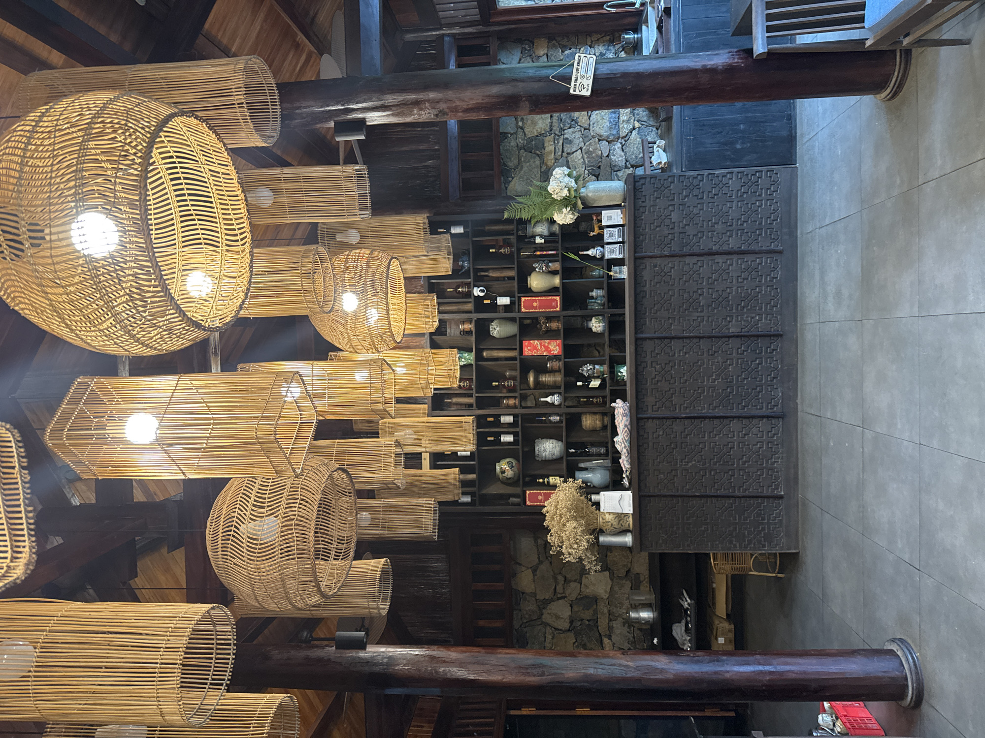 This image depicts the interior of a reception or bar area, likely a cafe or restaurant, located in Moc Chau, Vietnam. The space is characterized by a rustic and natural aesthetic, with prominent use of bamboo and wood. Overhead, a collection of large, woven bamboo pendant lights hang, casting a warm glow. These fixtures vary in shape and size, contributing to a visually dynamic ceiling.
The background features a stone wall, adding to the natural, textured feel of the environment. In front of the stone wall is a dark, intricately carved wooden counter. This counter displays a variety of decorative objects, including ceramic vases, bottles, and dried floral arrangements. Shelving behind the counter holds more decorative items, bottles, and what appear to be small boxes.
On the left side of the counter, a large, dark wooden pillar stands, supporting the ceiling structure. A red plastic crate is visible near the base of this pillar. The floor is tiled with large, grey square tiles.
On the right side of the image, another dark wooden pillar is present, with a wooden chair and a small table partially visible. A sign reading "KINH CHAO QUY KHACH" (Welcome Guests) and "WIFI" is hanging from this pillar. Further to the right, a sink area is partially visible.
The overall impression is one of a welcoming and aesthetically pleasing space designed to evoke a sense of natural beauty and tranquility, commonly found in tourist destinations like Moc Chau, Vietnam. There are no people visible in the image.