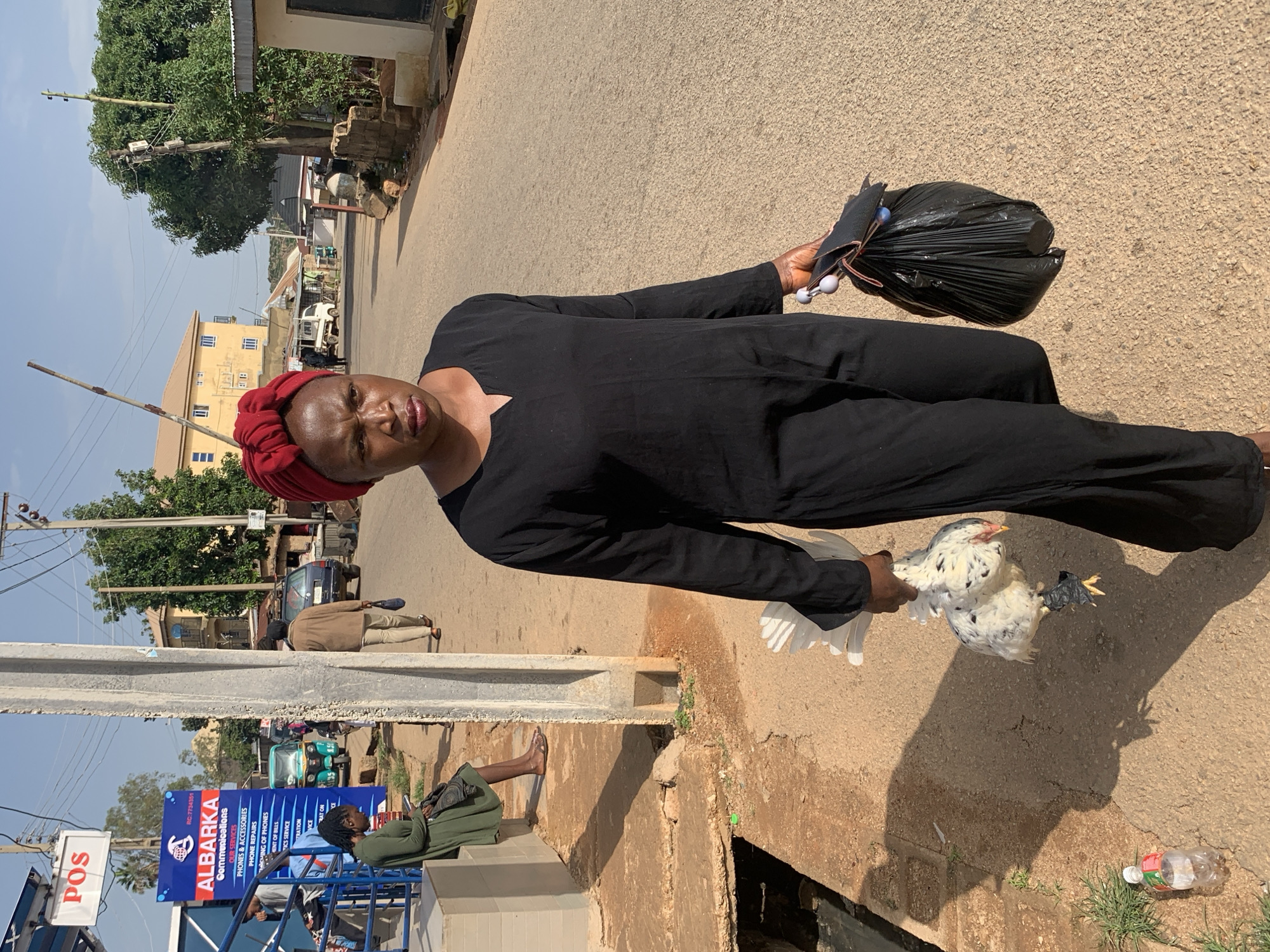 A woman wearing a black abaya and a red headwrap walks on a paved road in Gura Top, Nigeria. She carries a live white chicken in her right hand and a black plastic bag in her left hand. The chicken has black spots and its legs are bound with black tape. The woman is looking to her right with a serious expression. In the background, there are buildings, trees, and other people. One person is sitting on a ledge to the left, looking at a phone. Another person is walking away from the camera in the middle distance. A blue and white sign advertising "ALBARKA Communications" is visible on the left side of the image. The environment appears to be a commercial area with moderate pedestrian and vehicle traffic.