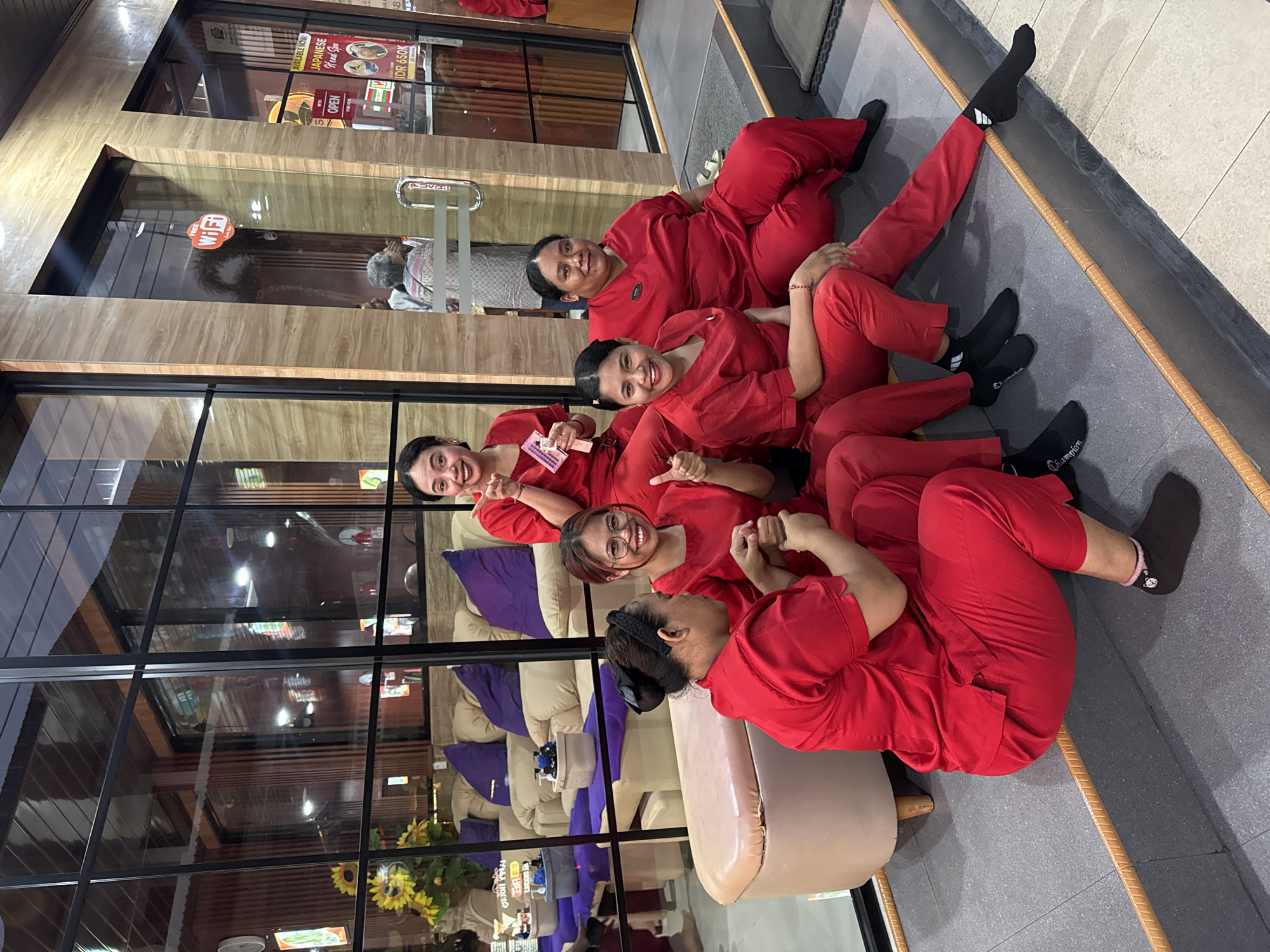 In Kuta, Indonesia, a group of five women, all wearing bright red uniforms, are posing for a photo outside a building with large glass windows. The building appears to be a spa or massage establishment, as indicated by signs visible through the windows advertising "JAPANESE HEAD SPA" and a price of "IDR 650K". The women are seated on grey steps in front of the building, with a beige bench to their left. Three of the women are in the foreground, with two in the middle and one on the far right. The woman on the far left is smiling and giving a thumbs-up. The woman next to her is wearing glasses and also smiling, giving a thumbs-up. The third woman in the foreground is also smiling. The two women in the background are also smiling. The environment suggests a warm, possibly tropical climate, with the building's facade featuring light-colored wood-grain paneling and a prominent "FREE WiFi" sticker on the glass door. Inside the building, plush sofas and tables are visible, hinting at a waiting area or treatment rooms. The lighting suggests it is either late afternoon or early evening.