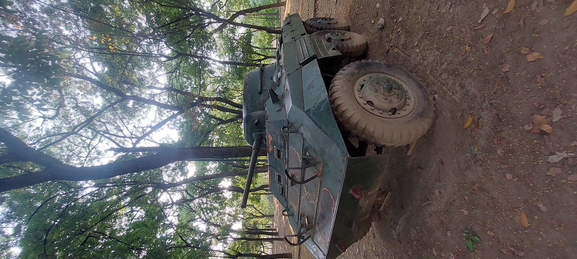 The image depicts an armored vehicle situated in a wooded area. The vehicle, possibly an older model armored car, is green with large, worn tires. The ground is covered in dirt and leaves, and trees surround the vehicle. The image appears to be taken outdoors during daylight. The city of Caracas, Venezuela is not evident from the image alone. The armored vehicle is resting on a dirt surface and is partially in shadow, with a mixture of leafy trees providing a background. There is no visible indication of people, actions, or interactions.