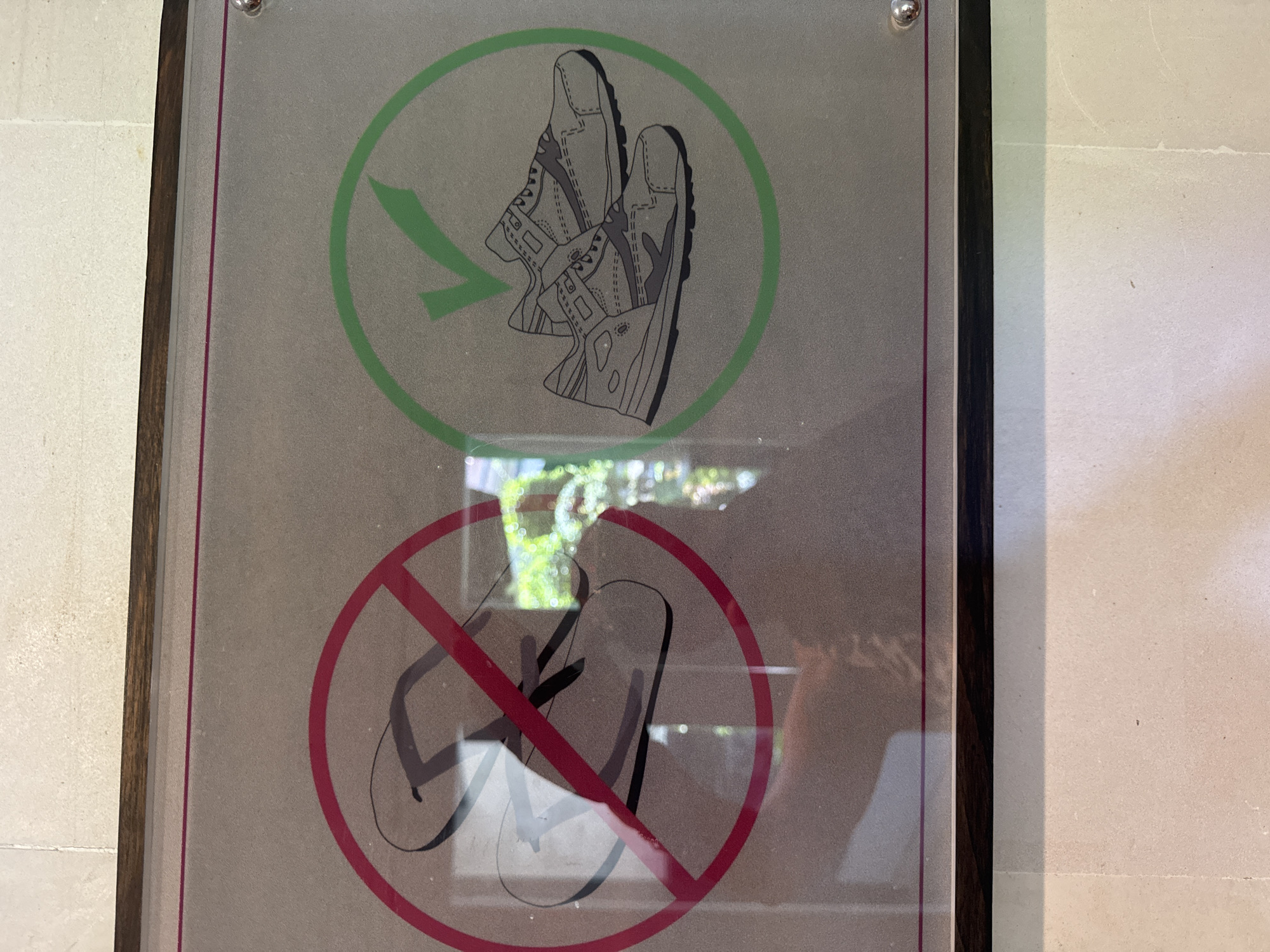The image shows a framed sign, likely located indoors on a light-colored wall, possibly in Kuta Selatan, Indonesia. The sign depicts two symbols: the top symbol shows two shoes inside a green circle with a green arrow pointing to the shoes, indicating that shoes are permitted. The bottom symbol shows two flip-flops inside a red circle with a red line crossing through them, indicating that flip-flops are prohibited.