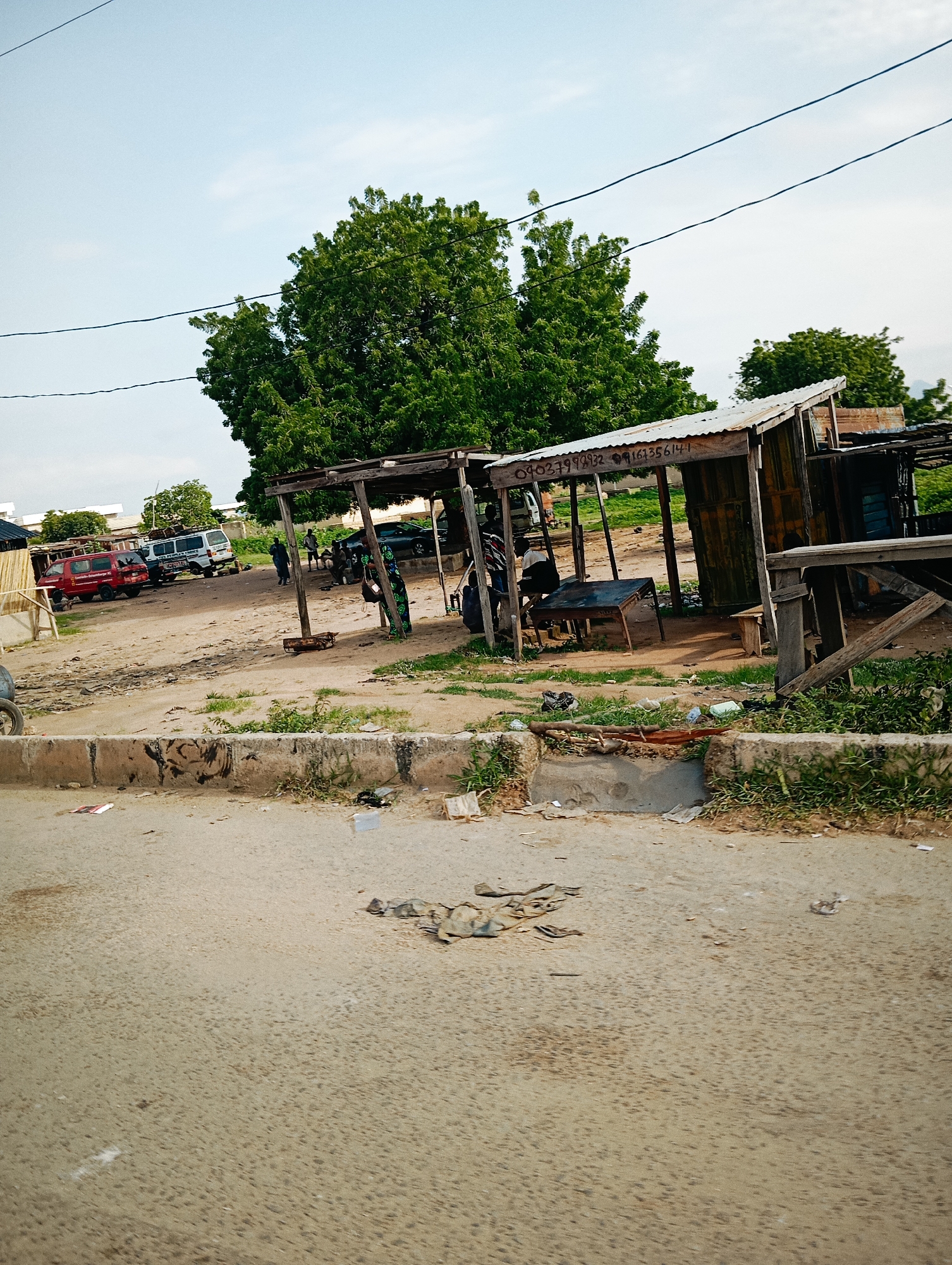 In Jalingo, Nigeria, a scene unfolds near a roadside shelter constructed with a corrugated metal roof supported by wooden posts. The shelter houses a table and chairs, suggesting a place for rest or informal business. In front of the shelter, a wooden beam extends horizontally, possibly part of the structure or an improvised bench. To the left, a red SUV and a white van are parked on a dirt lot, with a few individuals visible near the vehicles. Beyond the parked cars, more trees and structures dot the landscape. The foreground is dominated by a concrete curb and a dusty road, with scattered debris, including plastic bags and paper, littering the ground. The overall impression is of a utilitarian, somewhat disorganized, roadside stop in a developing urban environment.