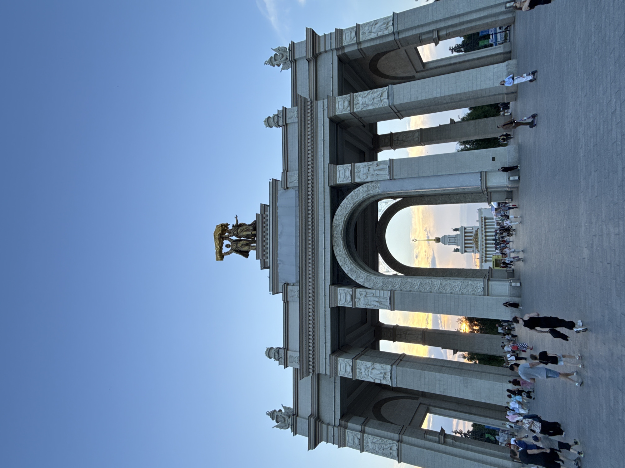 The image shows the Main Arch of the Exhibition of Achievements of National Economy (VDNKh) in Moscow, Russia. The arch is a monumental structure made of white stone, featuring multiple arches and intricate sculptures. Atop the arch, a gilded sculpture of two figures holding a wreath of wheat is visible. The sky is clear blue with some wisps of clouds, indicating it is likely late afternoon or early evening.
In the foreground, a paved plaza is filled with people walking and interacting. Several individuals are walking in various directions, some alone, others in groups. Children are also present, some walking with adults. A few people are riding electric scooters.
Through the arches of the Main Arch, a grand building with a spire is visible in the distance, also illuminated by the setting sun, creating a warm glow. Trees and greenery can be seen around the distant building. The overall scene depicts a public space bustling with activity during a pleasant time of day.